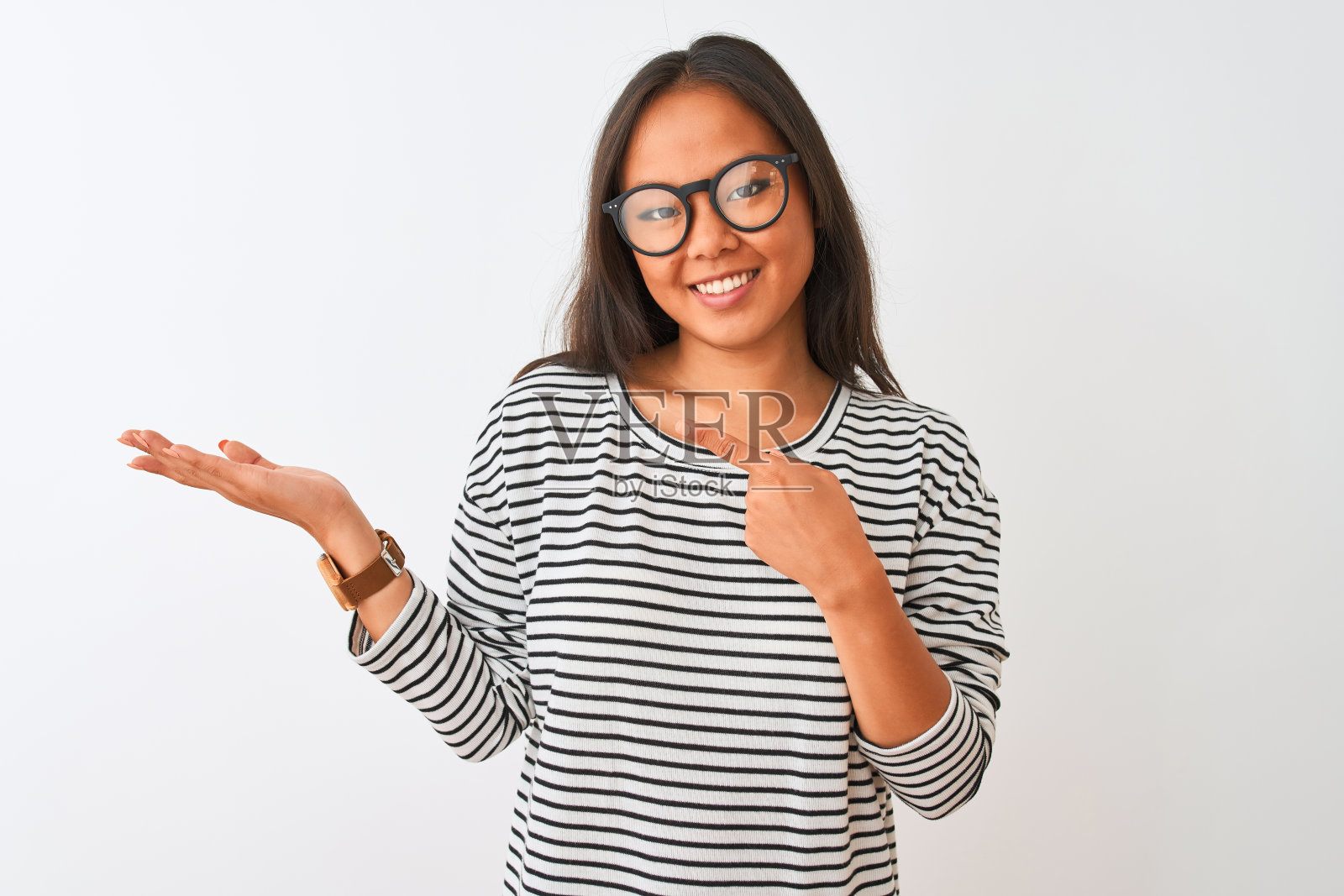 在孤立的白色背景下，一名年轻的中国女子身穿条纹t恤，戴着眼镜，一边用手指着镜头，一边惊讶地微笑着。照片摄影图片