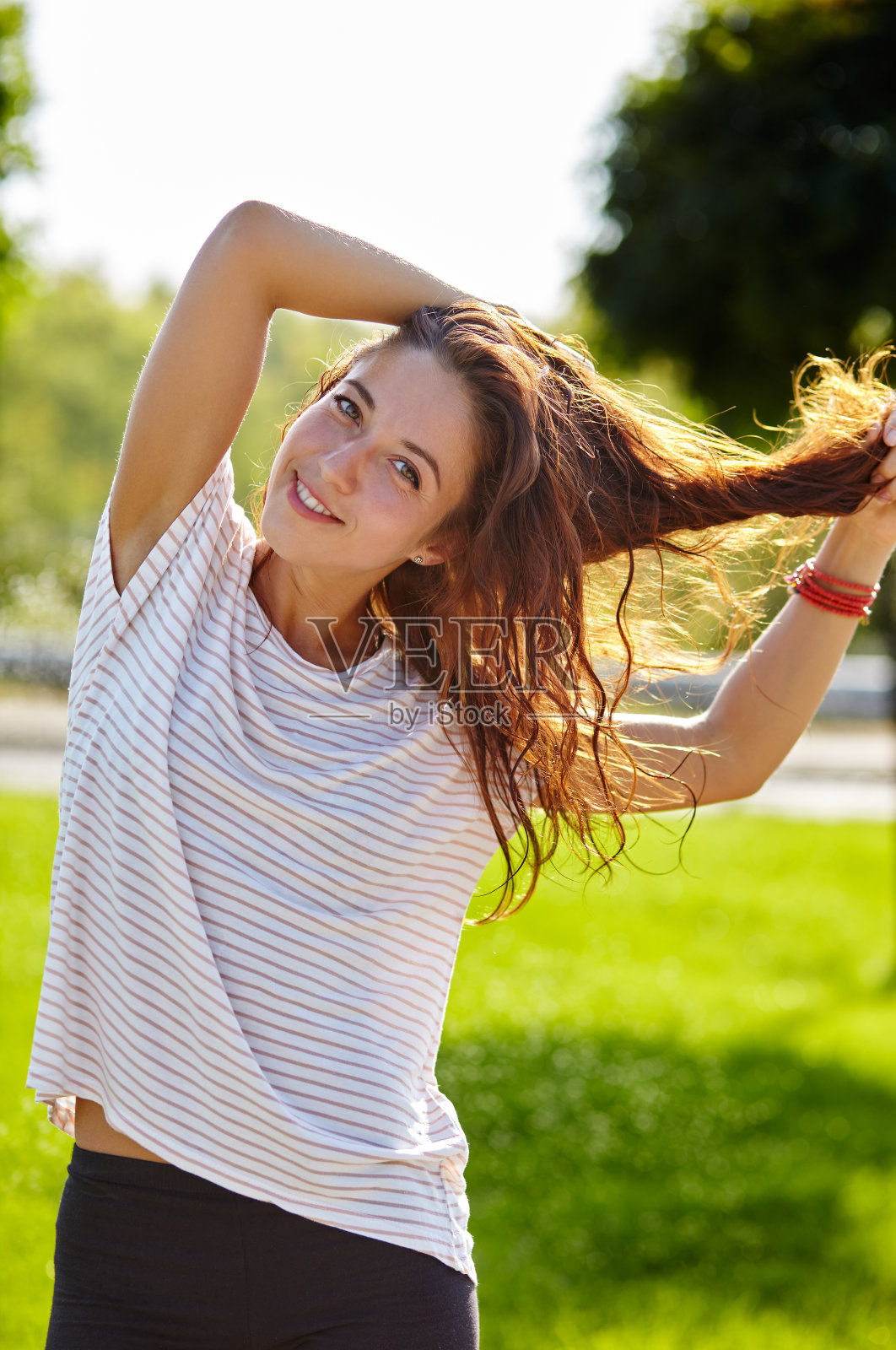 美丽迷人的女人穿着条纹t恤在夏天的公园摆姿势。活泼微笑的女孩手里拿着头发照片摄影图片
