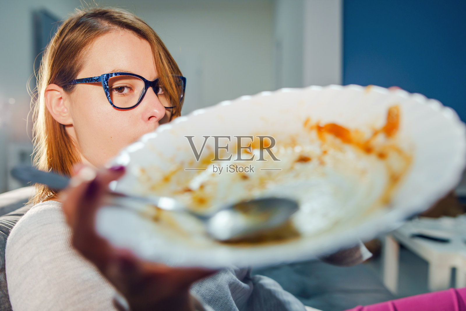 年轻的白人妇女坐在家里的床沙发上吃意大利面午餐或晚餐，穿着毛衣和眼镜，拿着白色的空盘子和勺子照片摄影图片