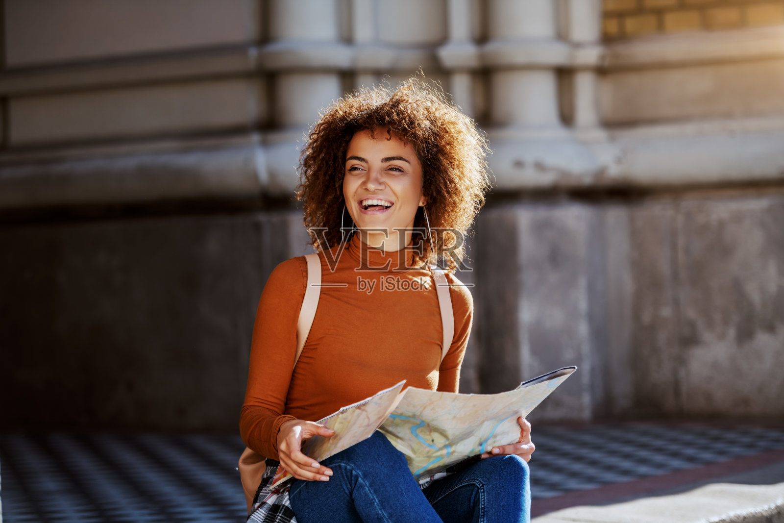 美丽的年轻混血女游客卷头发坐在城市街道上，拿着地图。照片摄影图片