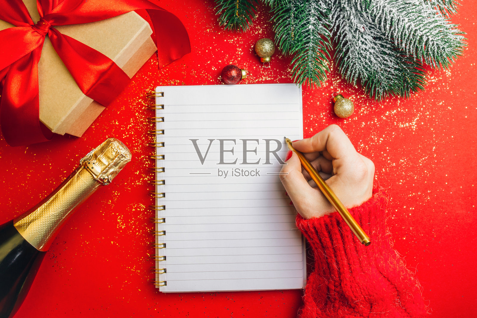圣诞礼物，香槟瓶，空白笔记本和杉树枝在红色闪闪发光的背景。女人用手在笔记本上写下新年目标或计划。平躺，有空间给你问候照片摄影图片