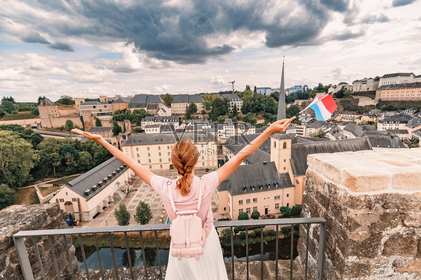 一个快乐的亚洲旅行者女孩举着卢森堡的旗帜，从观景台欣赏Grund区域。乡村旅游，娱乐和生活。照片摄影图片