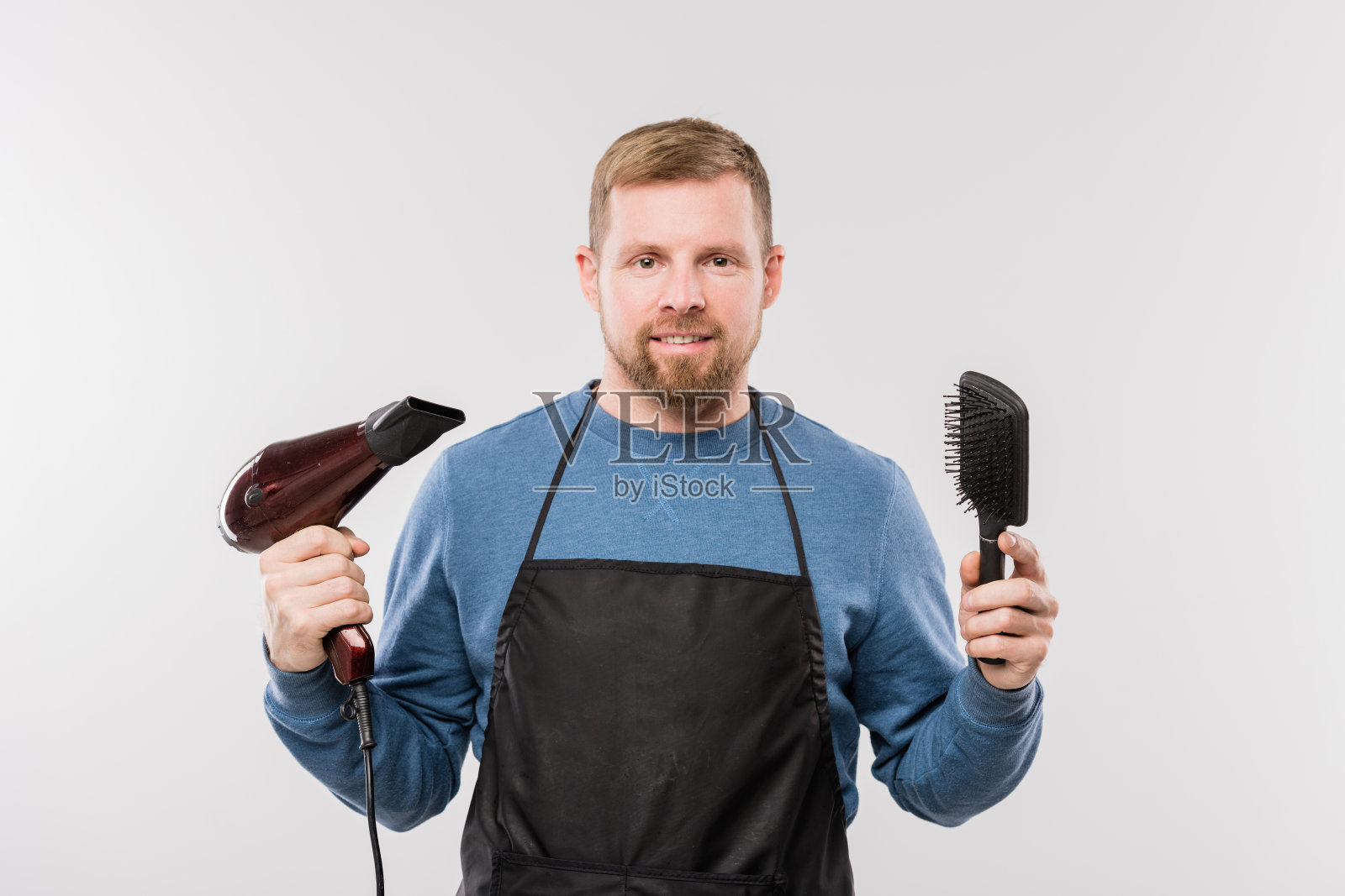 快乐的年轻胡子理发师在围裙拿着吹风机和发刷照片摄影图片