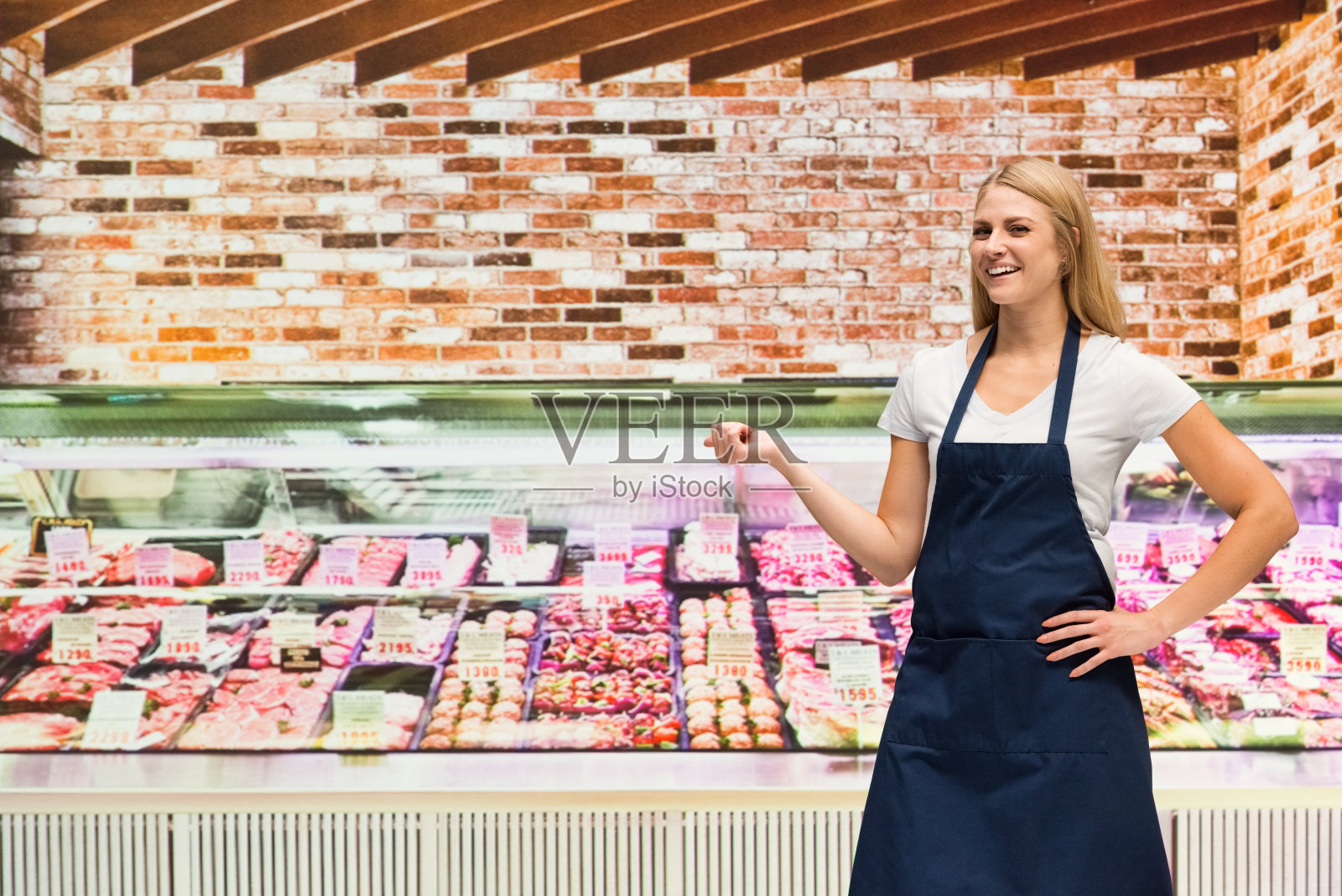 一个人/腰部以上20-29岁的成年人漂亮的金发/长发白人女性/年轻女性屠夫/店主站在肉店/熟食店/超市/市场-零售空间/商店的冷藏区照片摄影图片