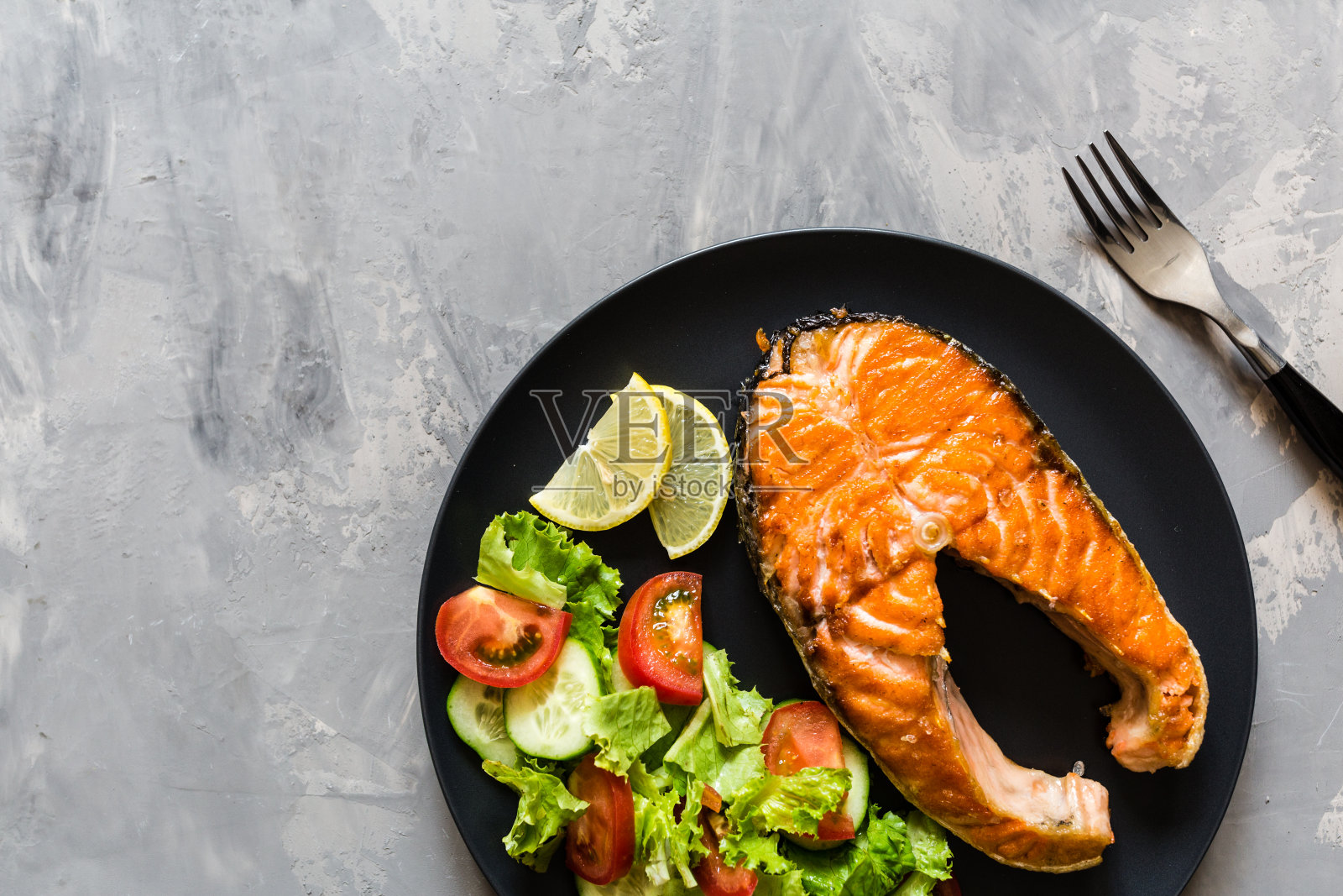 煎鲑鱼排配蔬菜沙拉和柠檬在黑盘子里。创意的布局由鱼，生菜，番茄，黄瓜，俯视图，平铺，模型，头顶。健康食品的概念照片摄影图片