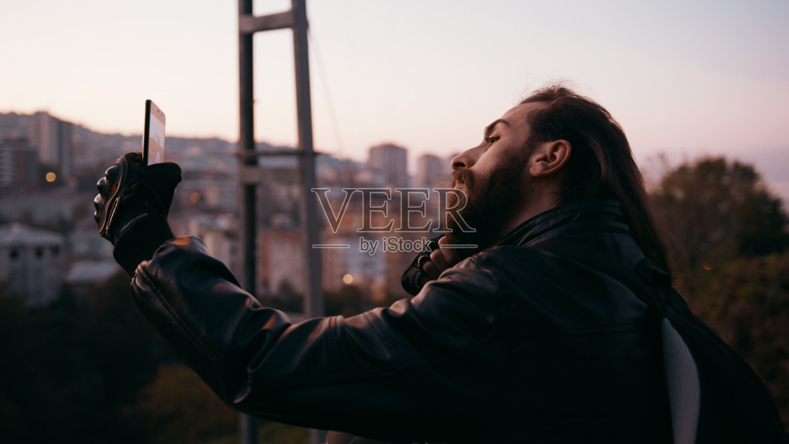 一名留着长胡子的年轻潮男正在以城市景观为背景自拍。照片摄影图片