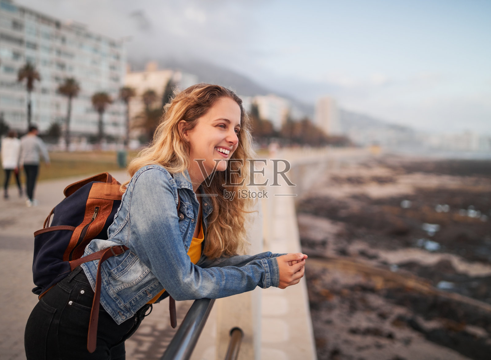 一个微笑的女性旅行者与她的背包在他的背部靠在栏杆上在城市街道望着别处的侧视图照片摄影图片