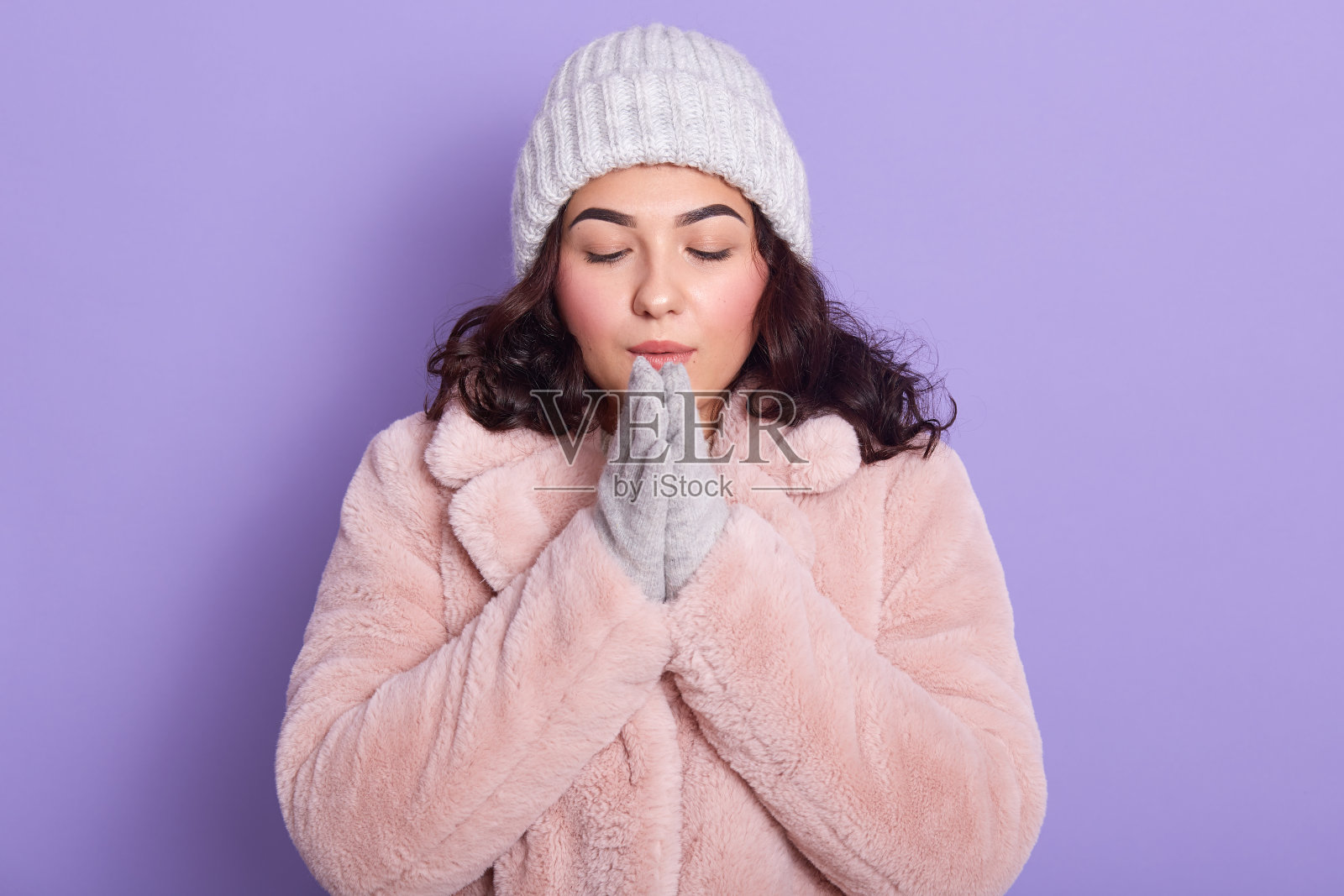 美丽的年轻女子在冬天晒太阳她的手，穿着暖和的衣服，人造皮大衣，手套和帽子，摆出孤立的丁香背景，闭着眼睛站着。冬季和寒冷天气概念。照片摄影图片