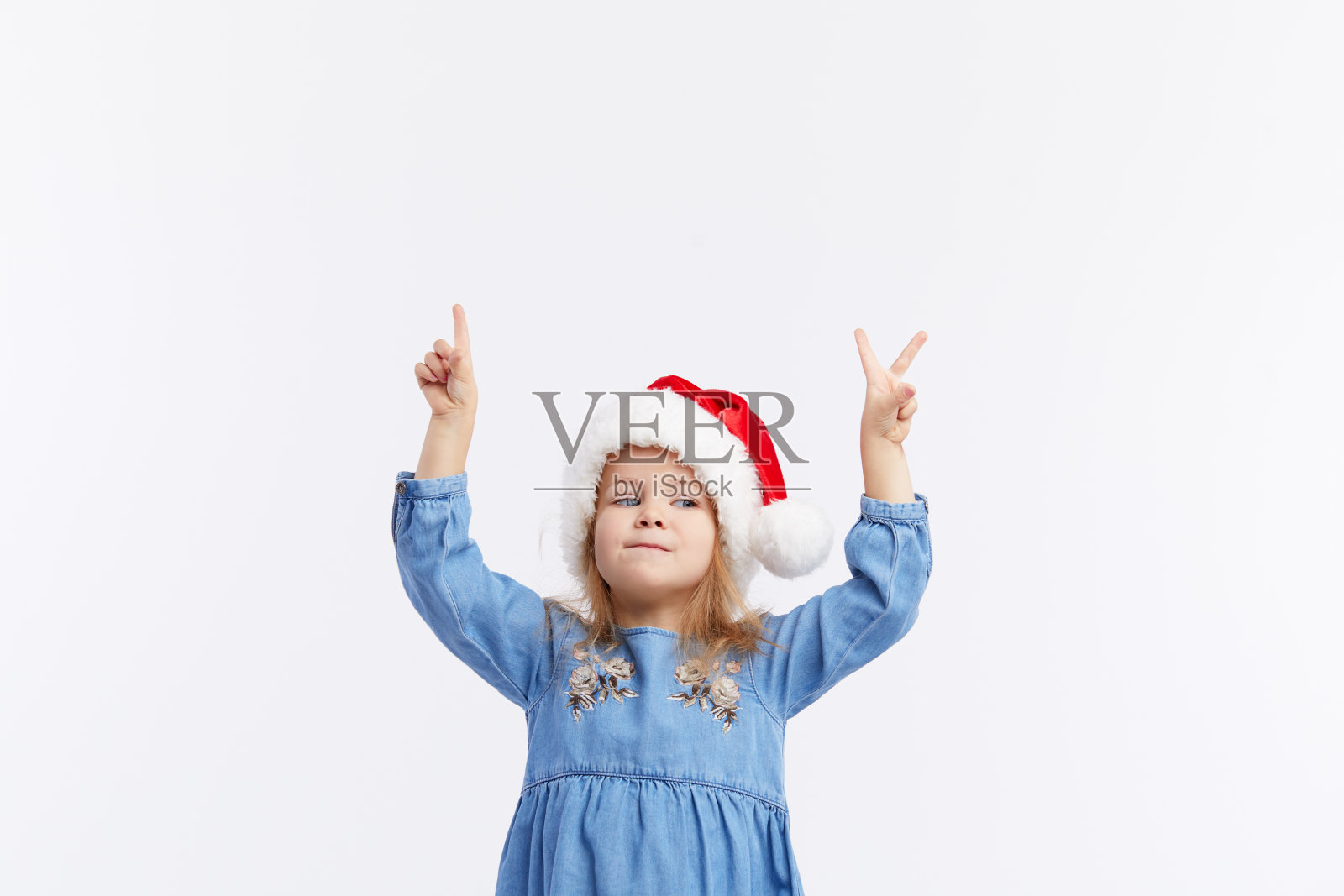 戴着圣诞帽和蓝色连衣裙的快乐微笑的女孩在红色背景上玩耍。指向上的手，文本空白区域。圣诞快乐，新年快乐照片摄影图片