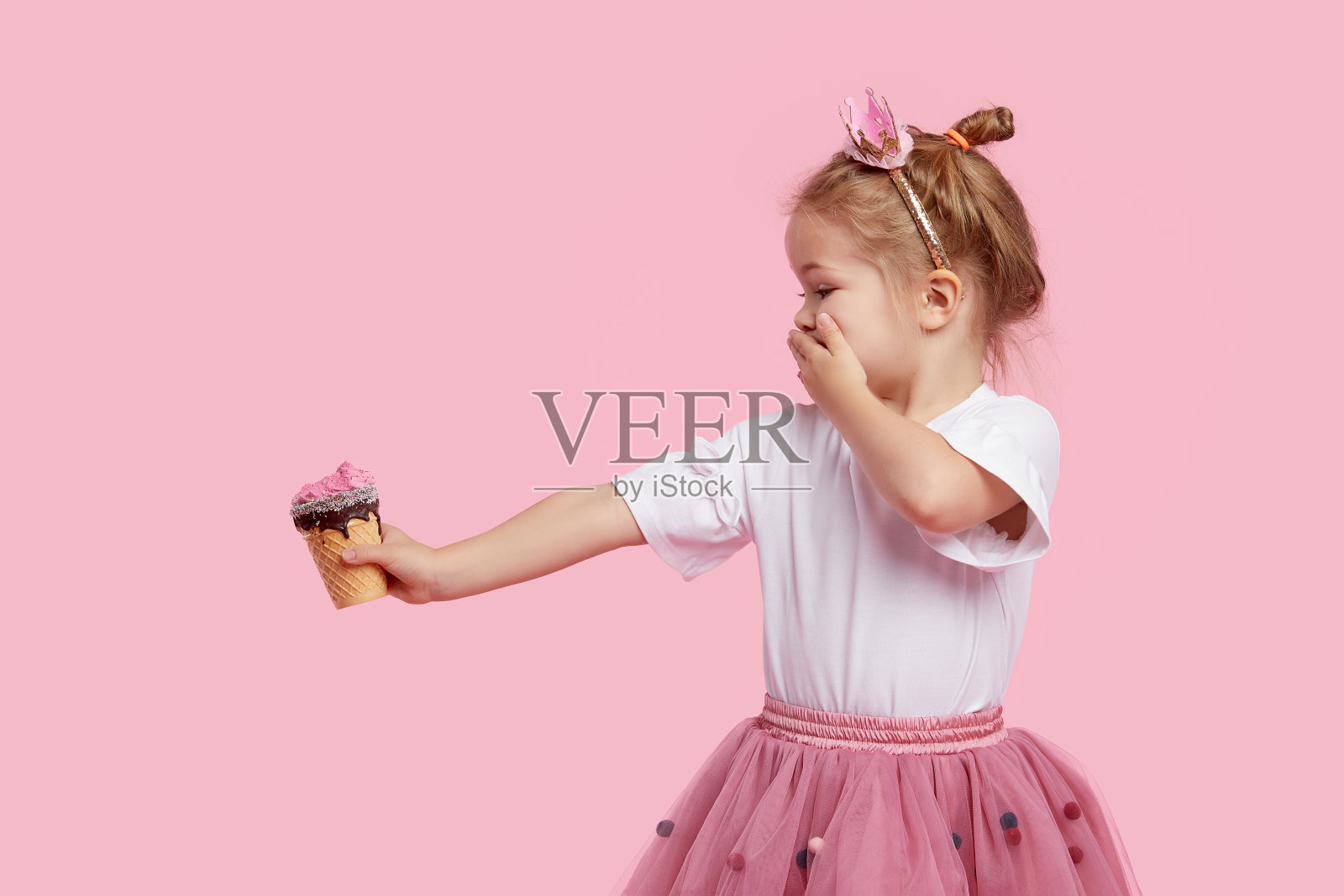可爱的小女孩把冰淇淋放在他伸出的手臂上，用手捂住他的嘴。在粉色的画室背景下拒绝吃无味的甜点。正确的婴儿食品不含糖照片摄影图片