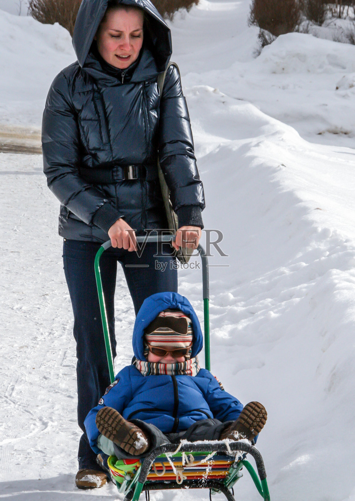 一位年轻的妇女推着她坐在雪橇上的小儿子。冬季娱乐。照片摄影图片