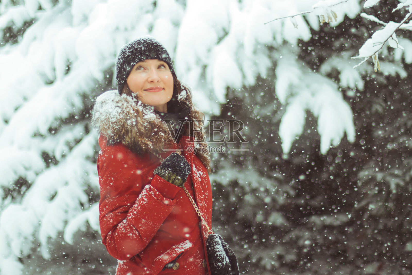年轻女子在红色羽绒服和黑色帽子仰望雪花，而外面是下雪-霜冻云杉树在雪的背景照片摄影图片
