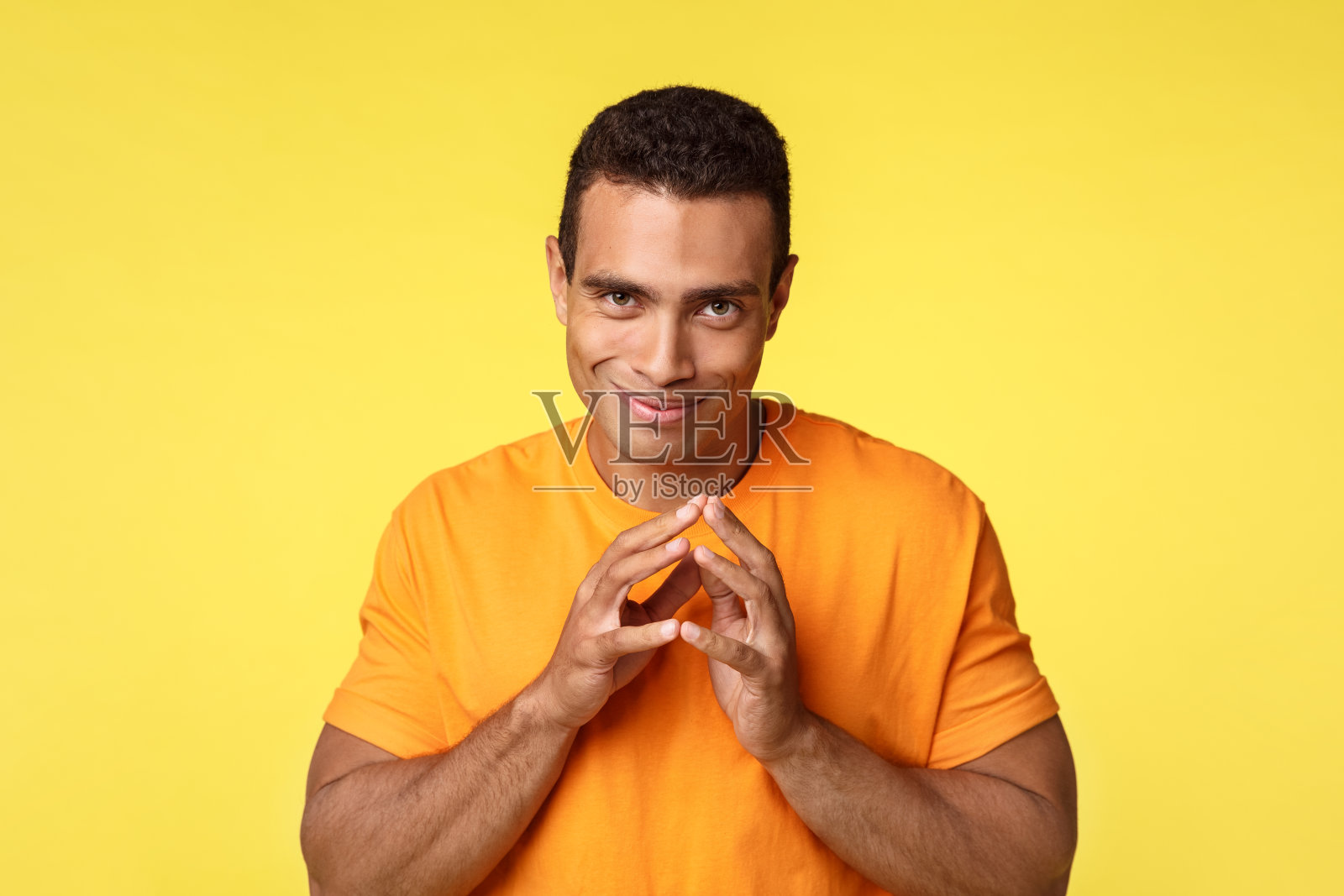 狡猾狡猾的年轻人有秘密的邪恶计划，傻笑着奸诈而尖尖的手指，准备复仇，站在黄色的背景快乐，想做坏事，穿橙色的t恤照片摄影图片