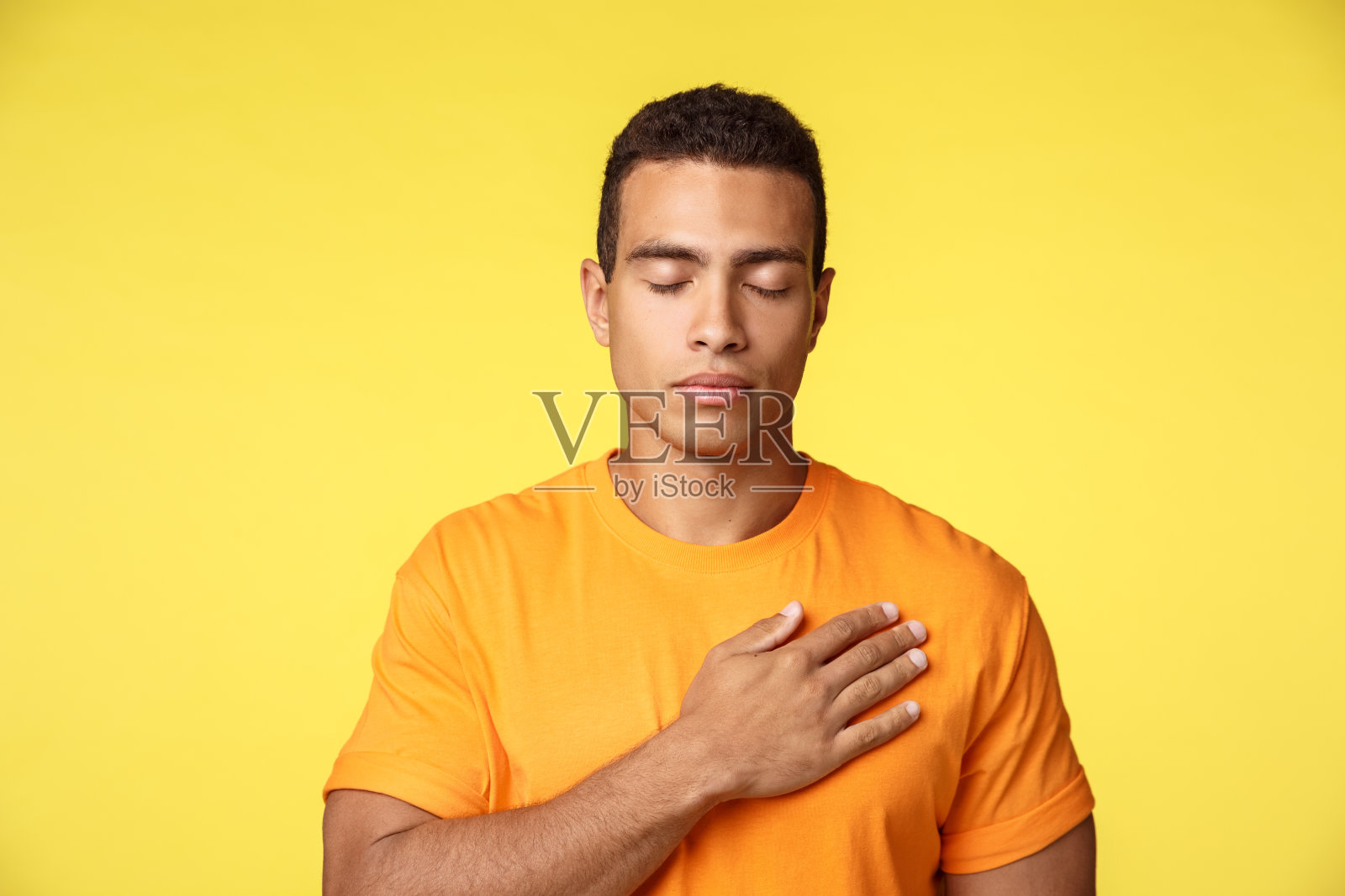 平静而充满希望的年轻帅哥把手放在心上，闭上眼睛祈祷或想着最亲爱的人，记住时刻，发誓，宣誓忠诚，站在黄色的背景照片摄影图片