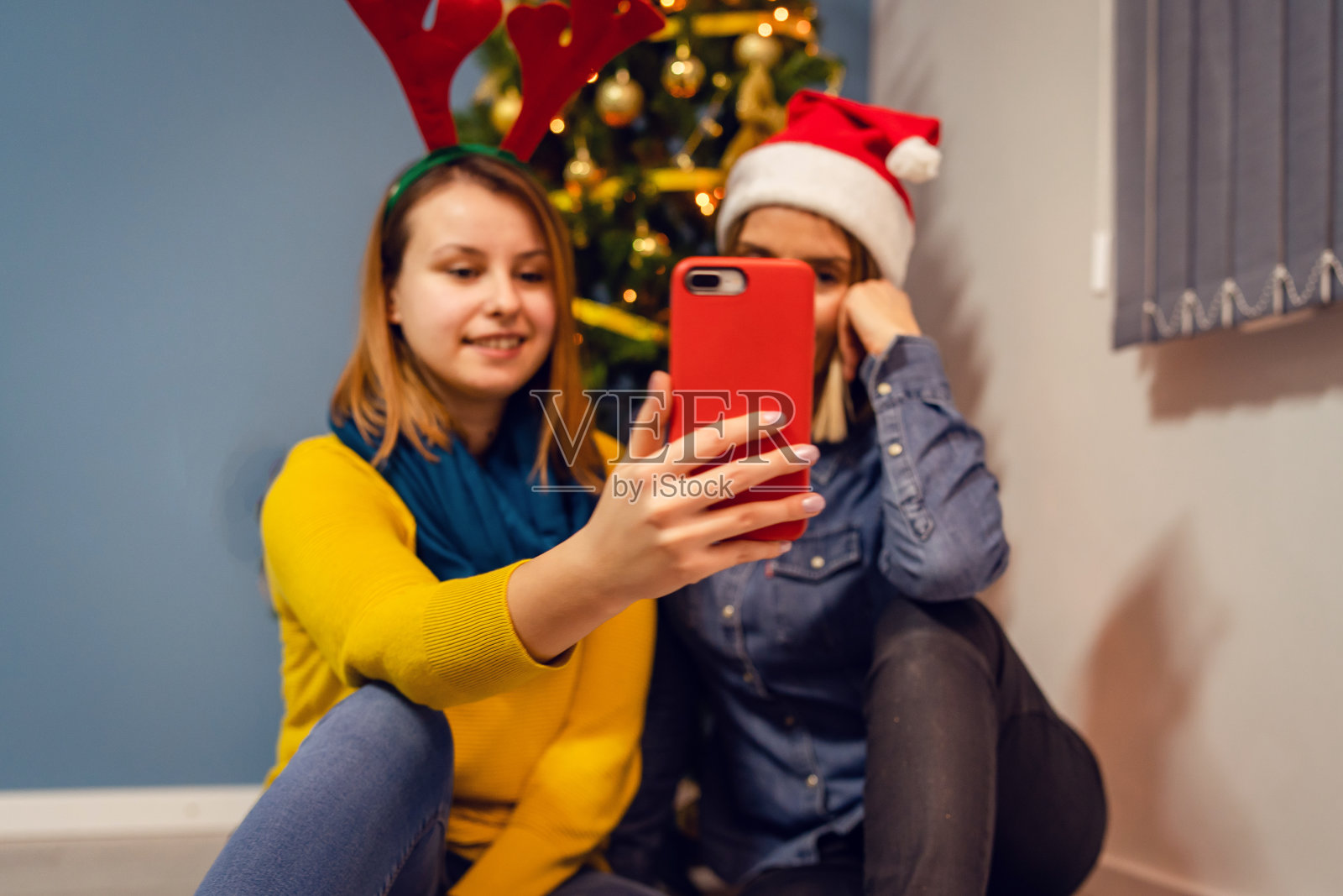 两位女性朋友女性或母女或姐妹坐在家里的圣诞树前用智能手机自拍视频聊天家庭圣诞帽照片摄影图片