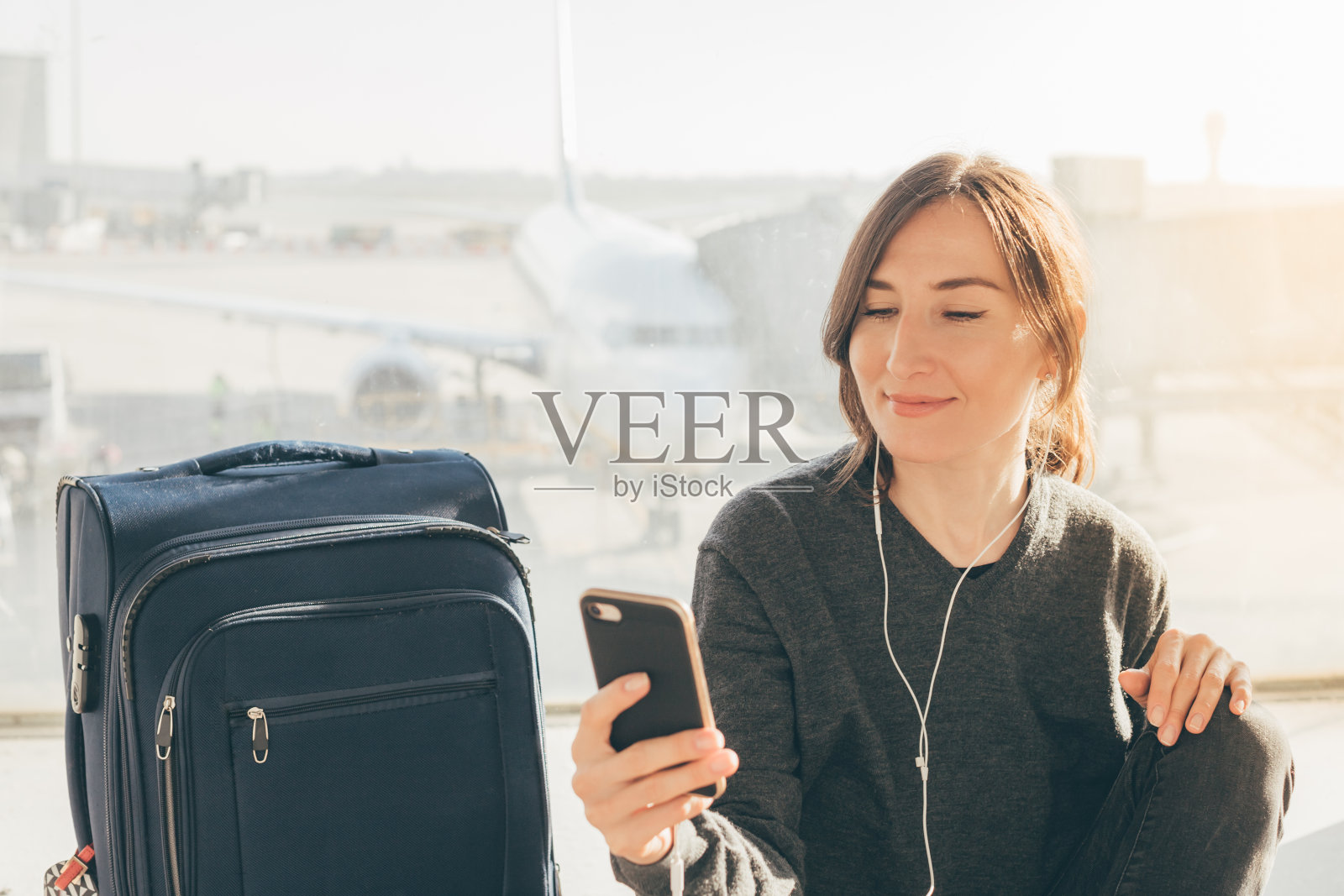 一名年轻女子带着她的手提行李和智能手机在机场候机厅等待她的航班(刚过了免税期)。丢失或取消航班-需要赔偿概念(巴塞罗那，西班牙)。照片摄影图片