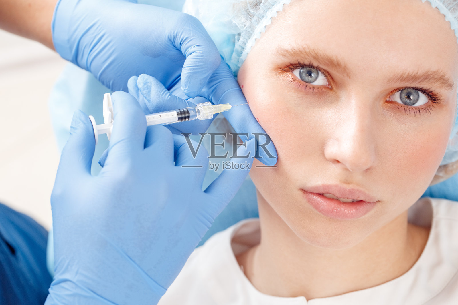 美容服务。年轻女子在美容诊所躺着看相机好奇，而医生在手套做注射透明质酸到脸颊特写照片摄影图片