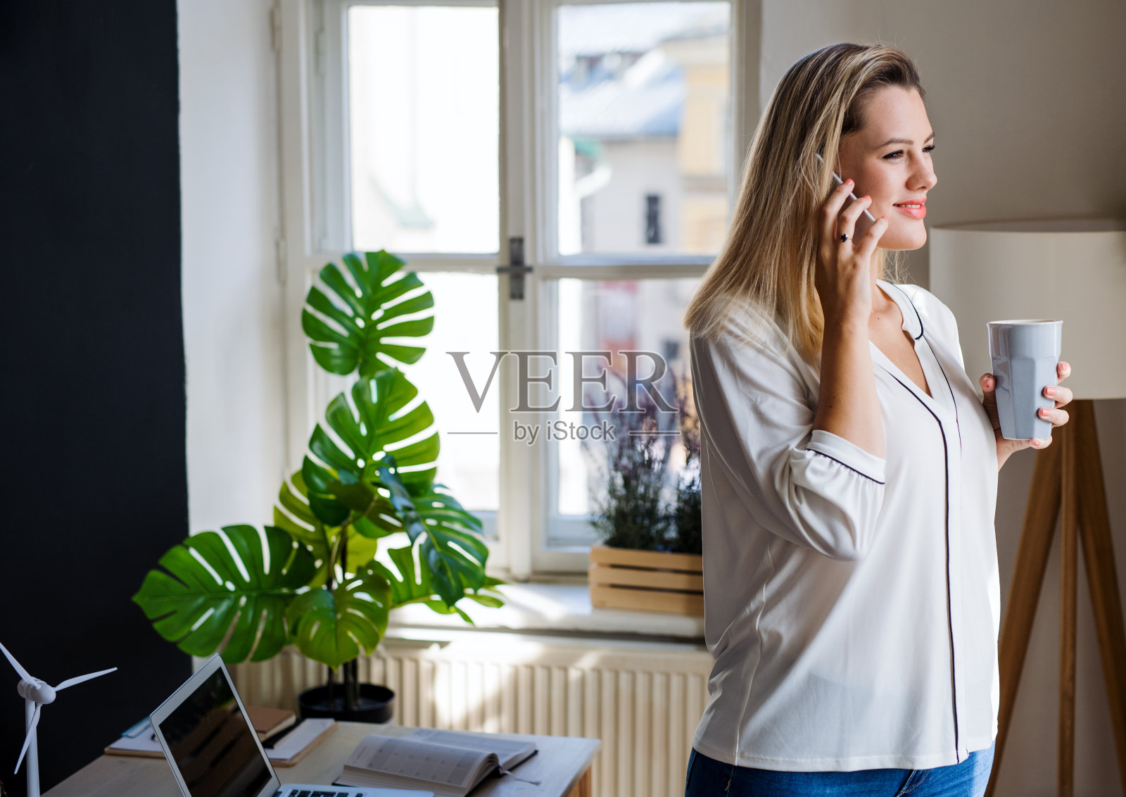 年轻女子拿着智能手机站在家里的办公室里工作。照片摄影图片