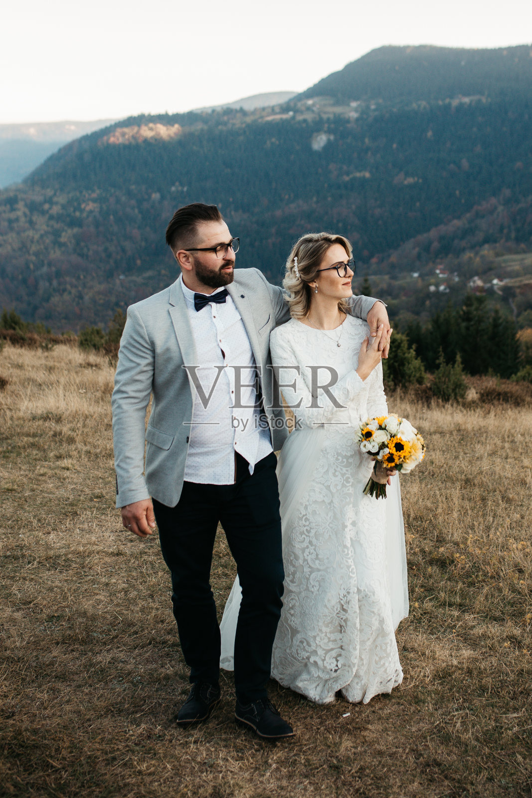 一对美丽的夫妇在他们的婚礼上有一个浪漫的时刻，在日落的山上。新娘身着白色婚纱，手里拿着一束向日葵，新郎身着西装。他吻了她。照片摄影图片