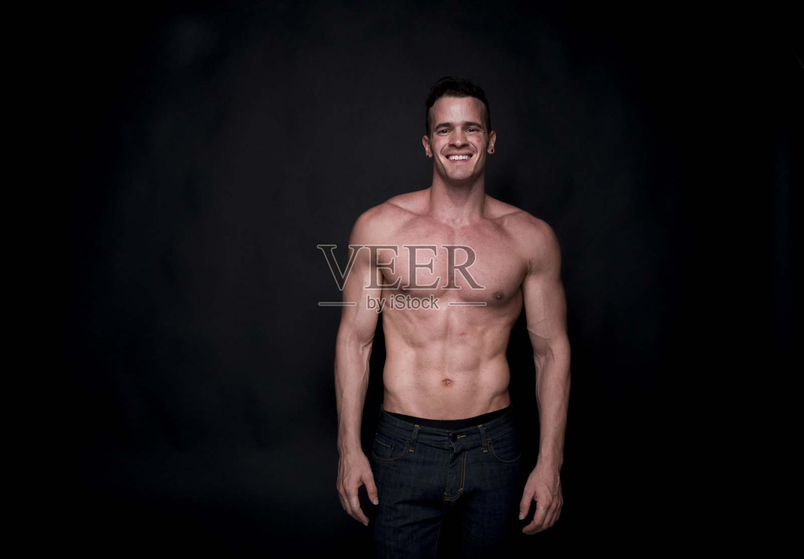 腰部以上/只有一个男人/一个20-29岁的成年人英俊的人白人男性/年轻男子站在黑色背景，穿着牛仔裤谁是肌肉发达/一个性感的象征/赤膊/黑色颜色照片摄影图片