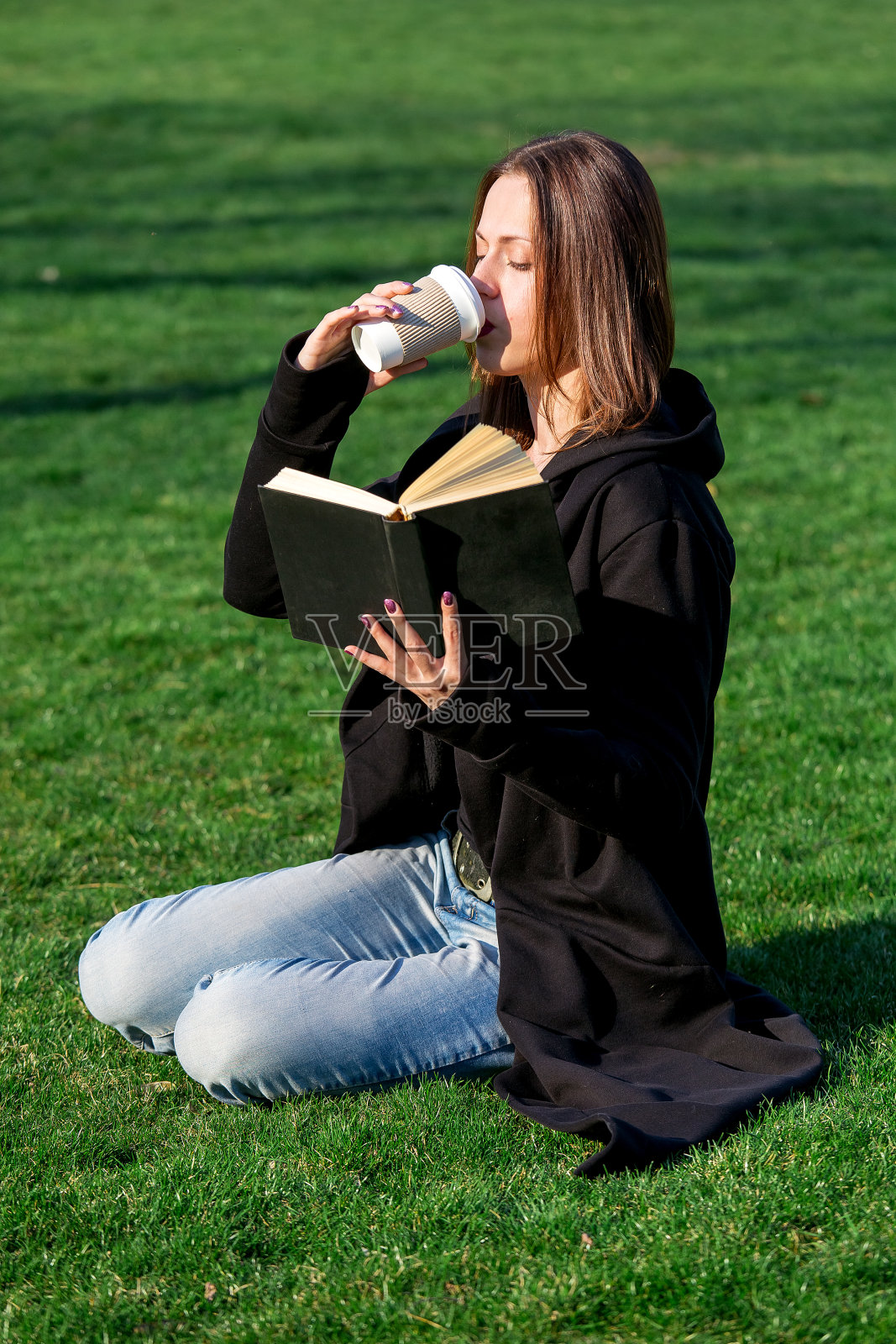一个黑发长发的女孩，穿着黑色的外套和蓝色的牛仔裤，坐在公园的草坪上，手里拿着一本书，喝着一次性纸堆里的热咖啡。照片摄影图片