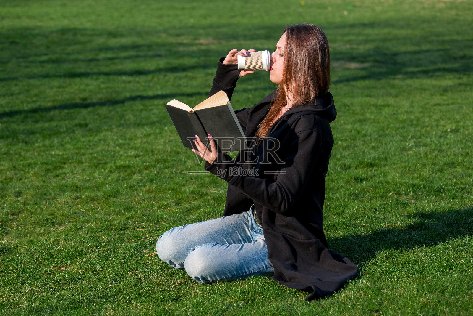 一个长头发的年轻女孩从一个纸杯喝咖啡，读一本黑色的精装书坐在绿色的草地上在一个阳光明媚的日子，侧视图。照片摄影图片