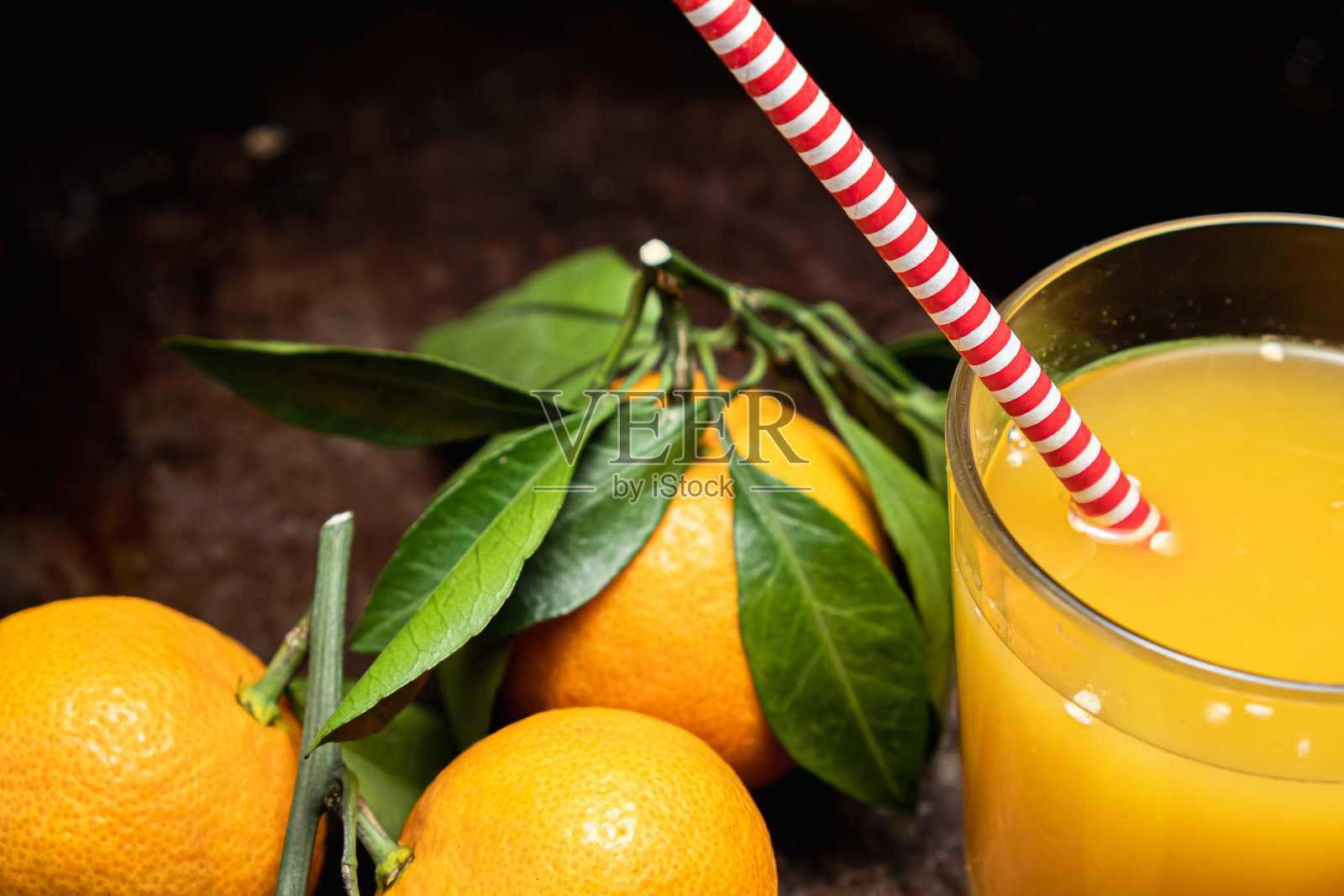 在锈迹斑斑的表面上，三个旁边带叶子的橘子是一个盛满果汁的玻璃杯。照片摄影图片