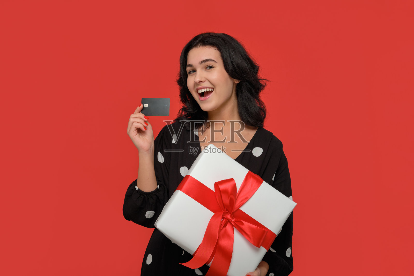 深色头发的女人穿着黑色的裙子微笑着拿着白色的圣诞礼物盒和银行卡。照片摄影图片