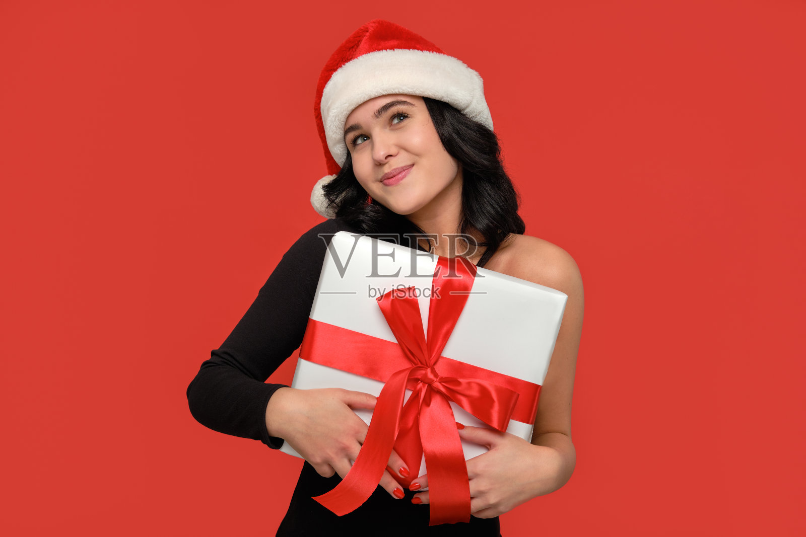 女人穿着一件连衣裙和圣诞老人的帽子微笑着拿着圣诞礼物盒白色的红丝带照片摄影图片