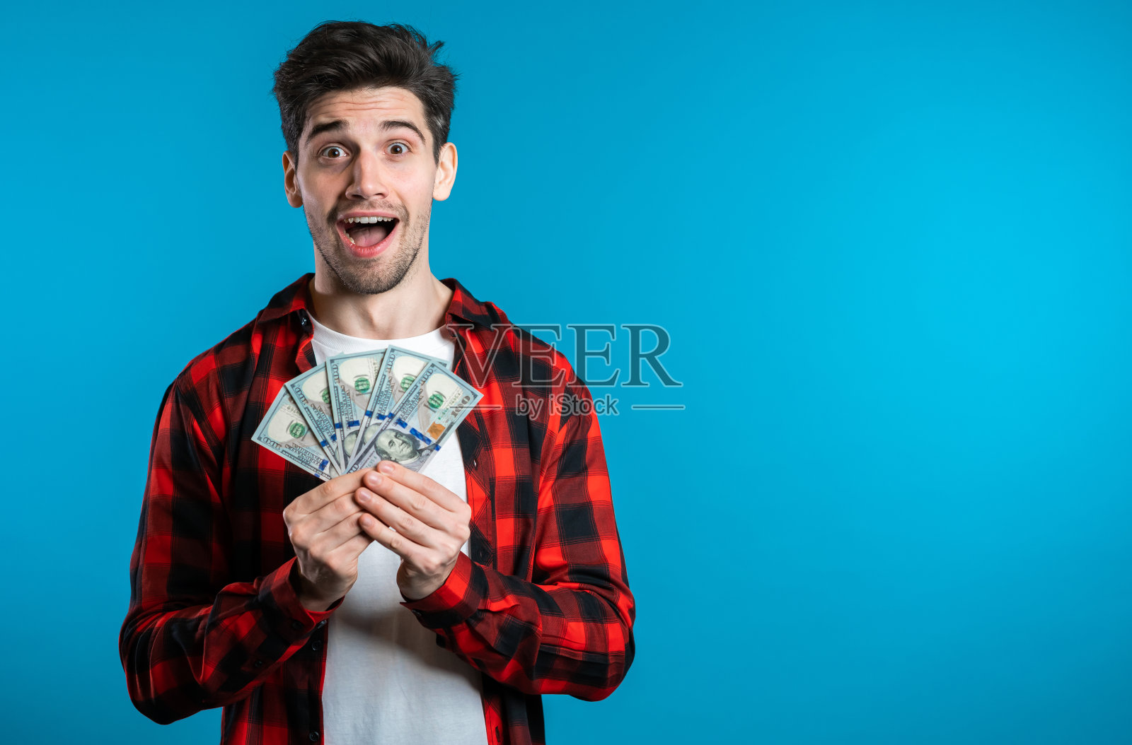 身着红色格子衬衫的男子，脸上带着惊讶的快乐表情，手里拿着美元。有金钱的人。新的美元在蓝色的工作室背景。照片摄影图片