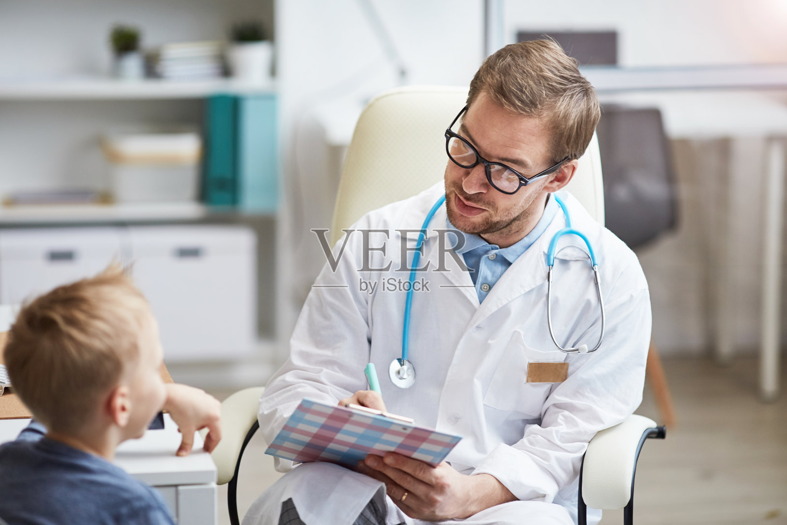 友善的年轻医生穿着白大褂与小患者交谈病情并在病历卡上做笔记照片摄影图片