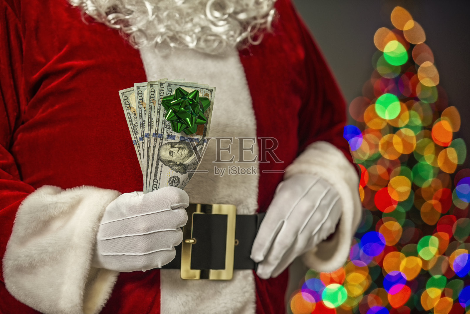圣诞老人拿着5张100美元的钞票，背景是圣诞树上的蝴蝶结和焦糖照片摄影图片