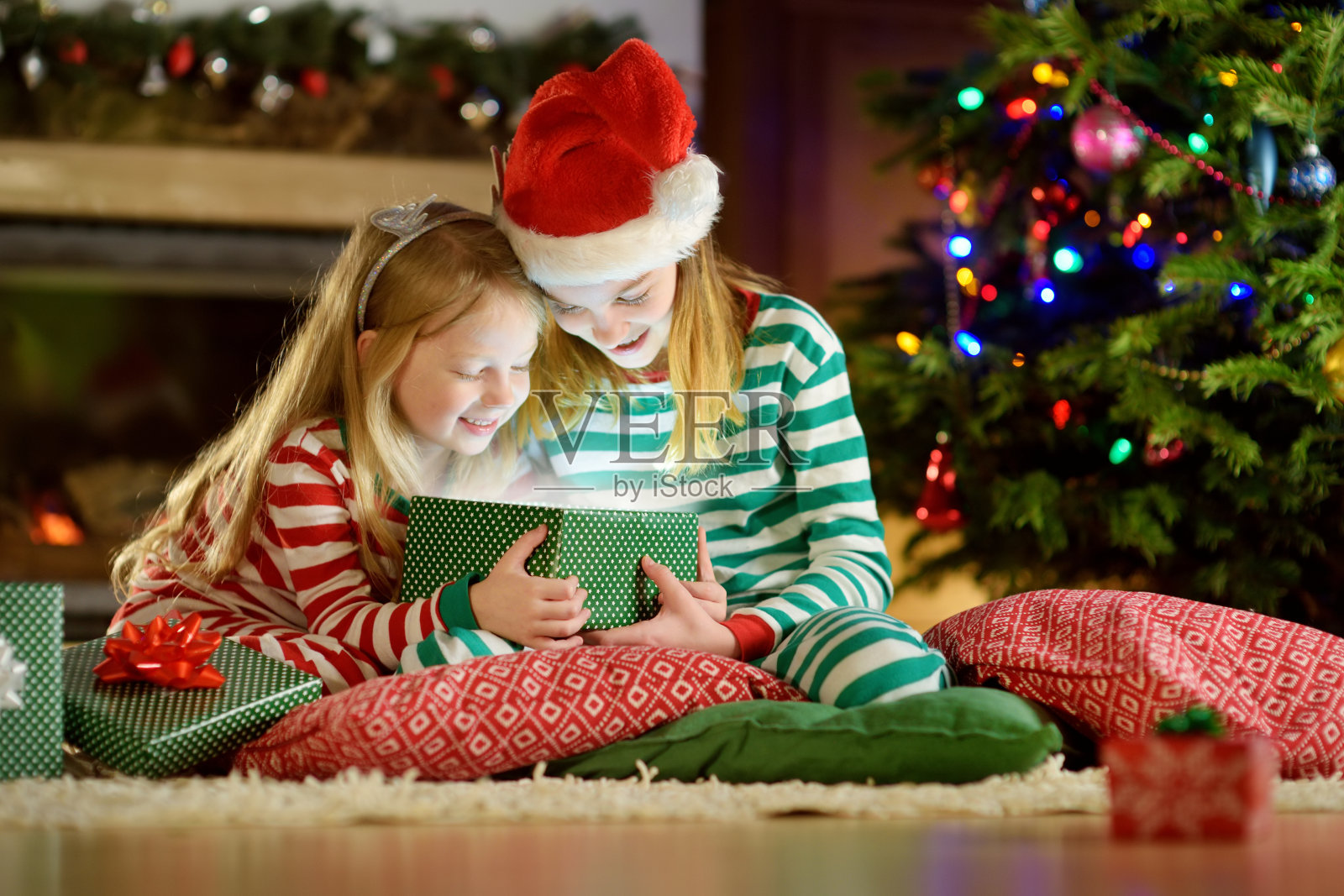 平安夜，快乐的小姐妹们穿着圣诞睡衣，在舒适黑暗的客厅壁炉旁打开礼品盒。照片摄影图片