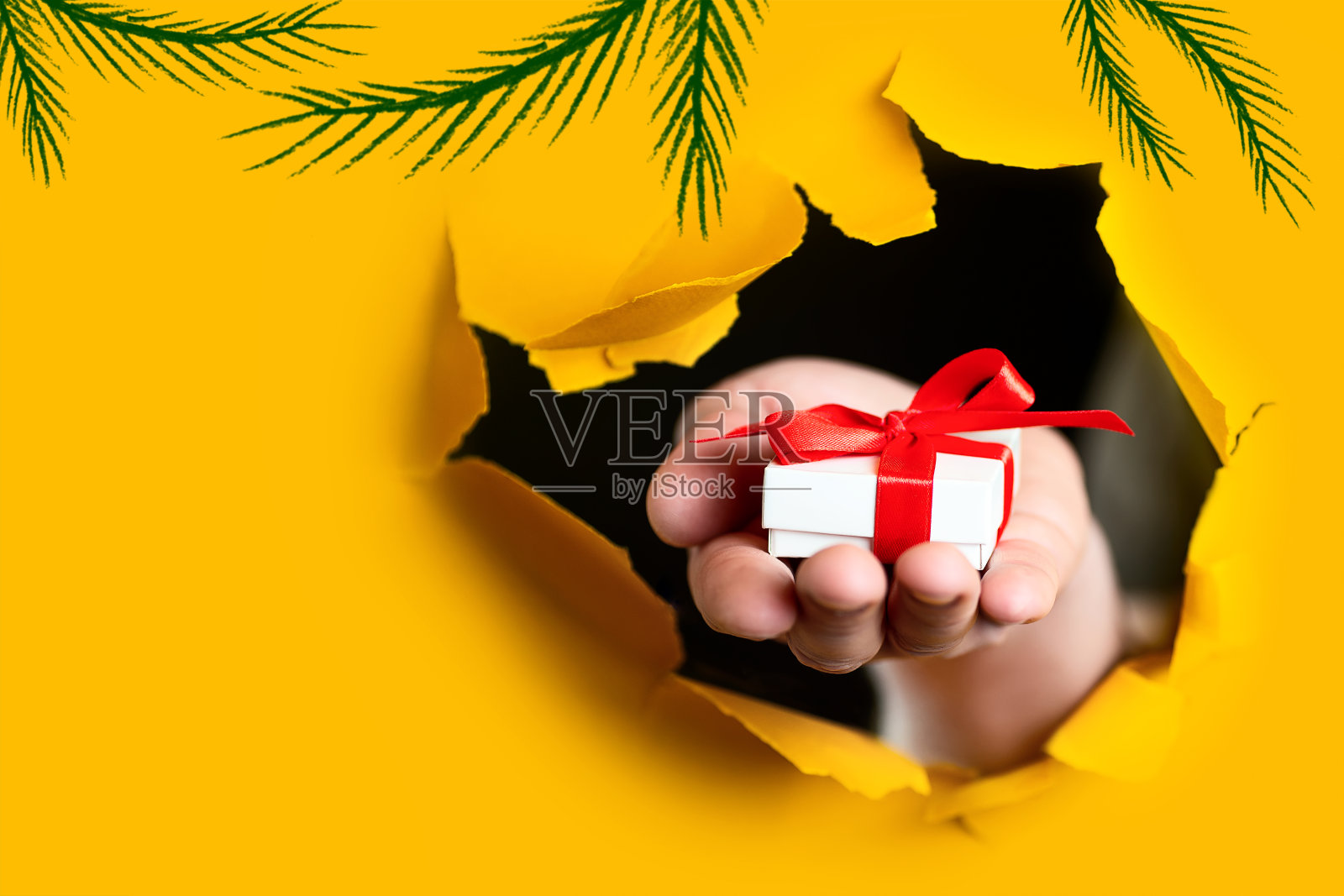 在黄色的纸背景下，一个破洞里露出一件礼物，手里拿着一个红色的蝴蝶结照片摄影图片