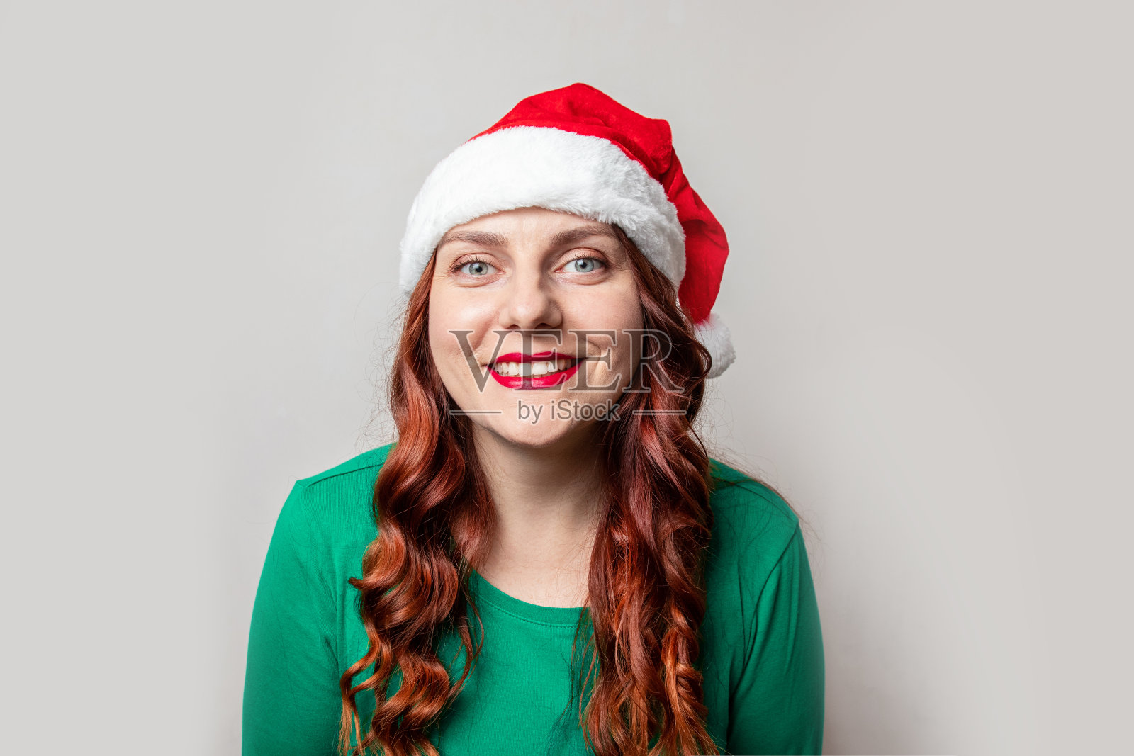 一个小女孩，一头红色的卷发，戴着一顶红色的圣诞帽，看着镜头，对着灰色的背景微笑照片摄影图片