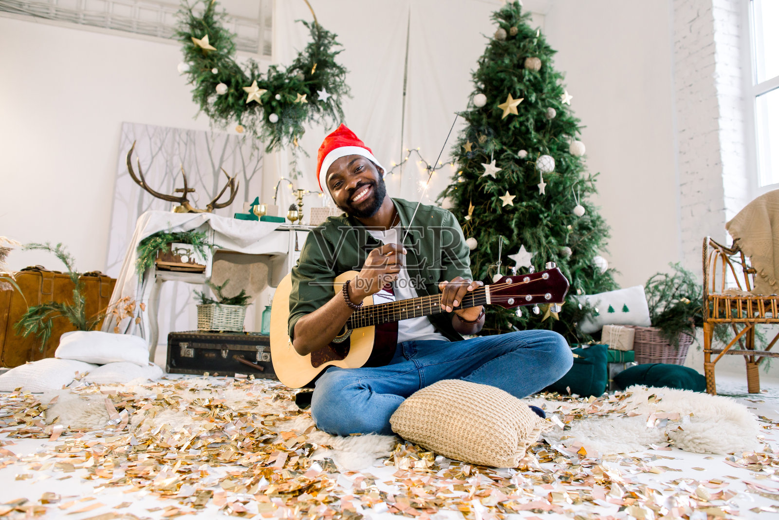 英俊的非裔美国男孩戴着圣诞帽，拿着吉他和孟加拉灯，坐在圣诞装饰品前的地板上照片摄影图片