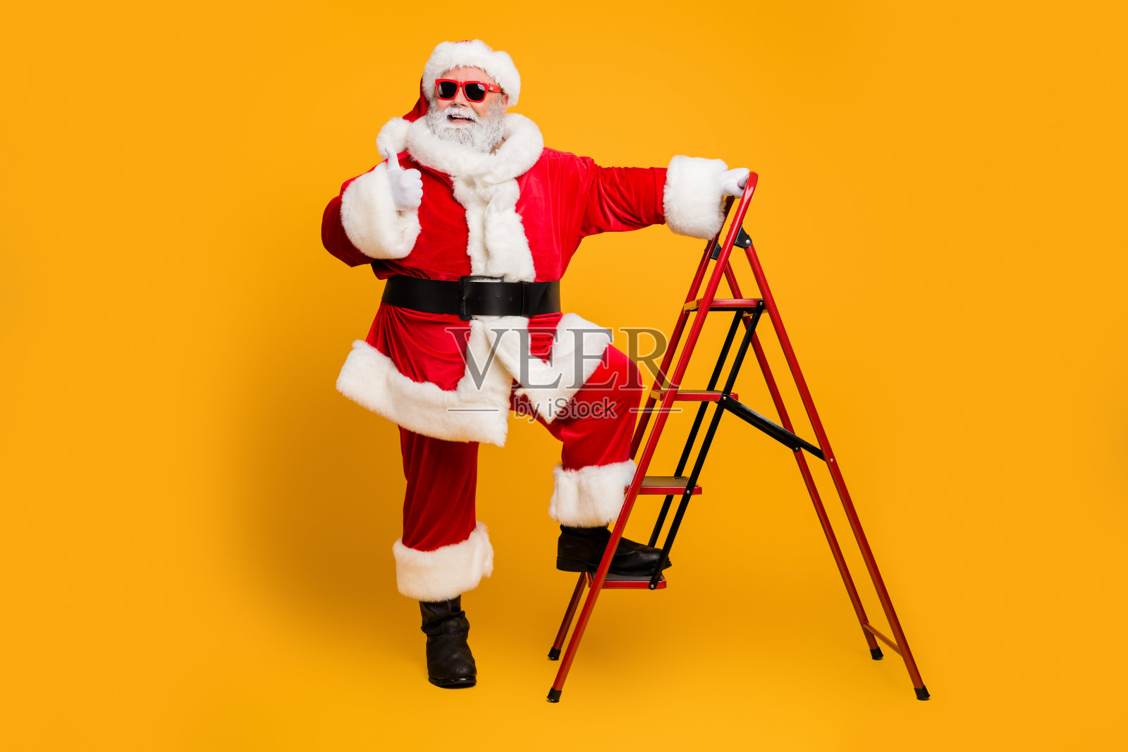 时髦的白胡子头发圣诞老人在红帽子爬梯子装饰圣诞树送祝愿梦想礼物显示拇指赞成圣诞节庆祝孤立的黄色背景照片摄影图片