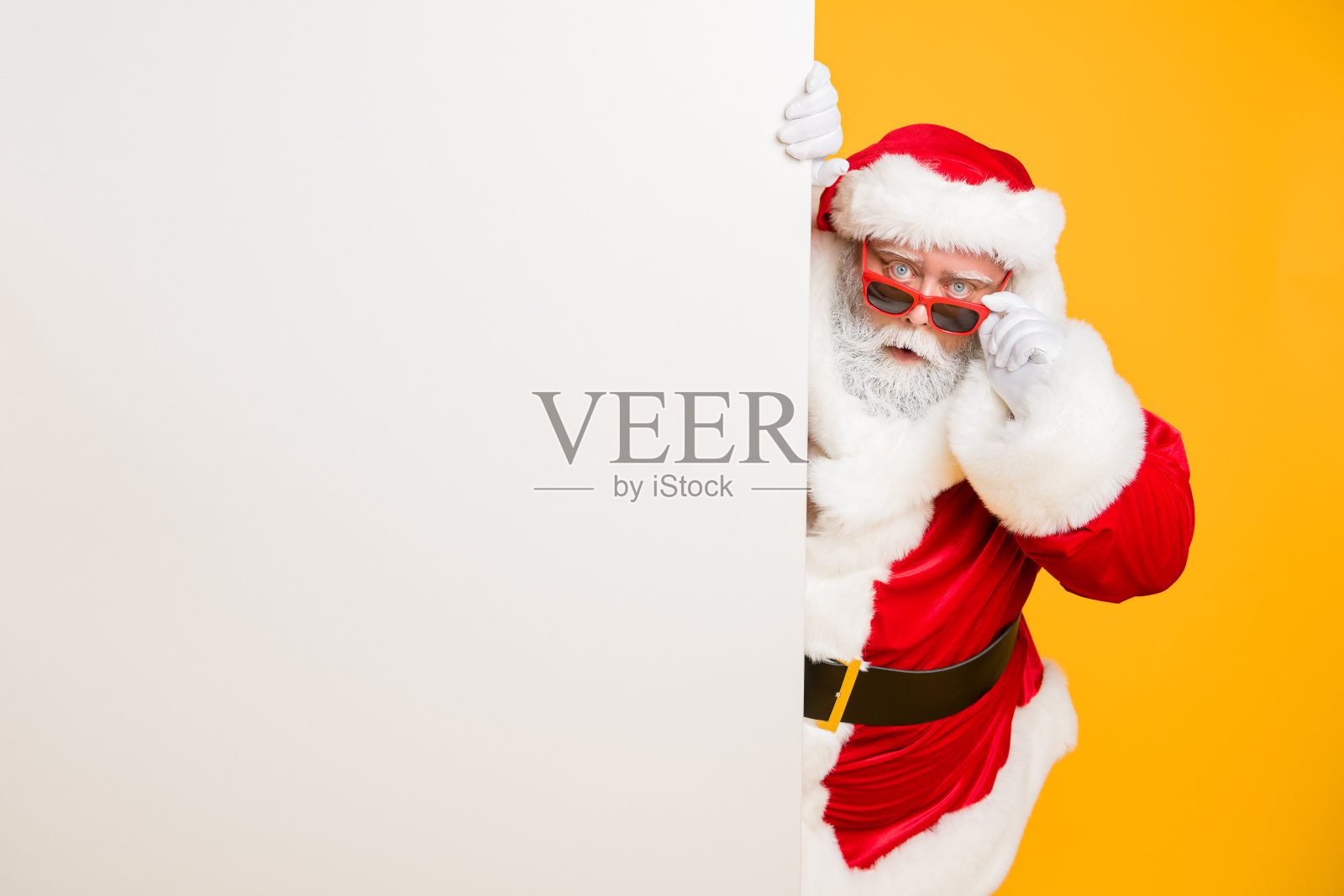 滑稽时髦的圣诞老人在红色头饰看后面的白色广告牌奇迹不可思议的2020年圣诞魔术事件穿着时尚的黑带孤立在黄色背景照片摄影图片