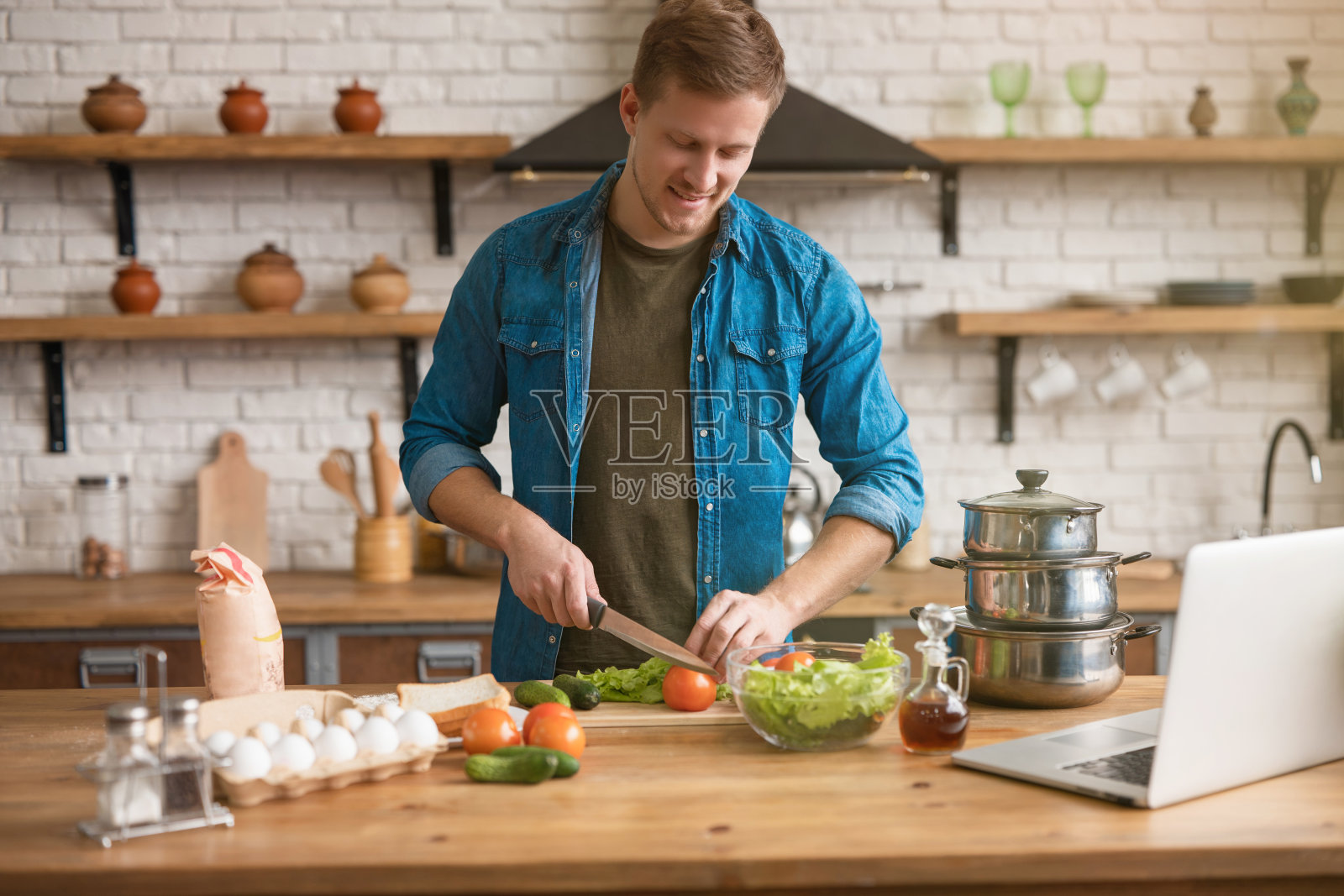 年轻英俊的男人在设备齐全的厨房里用刀和木盘切蔬菜，准备午餐沙拉，在笔记本电脑上查看食谱，周末氛围照片摄影图片