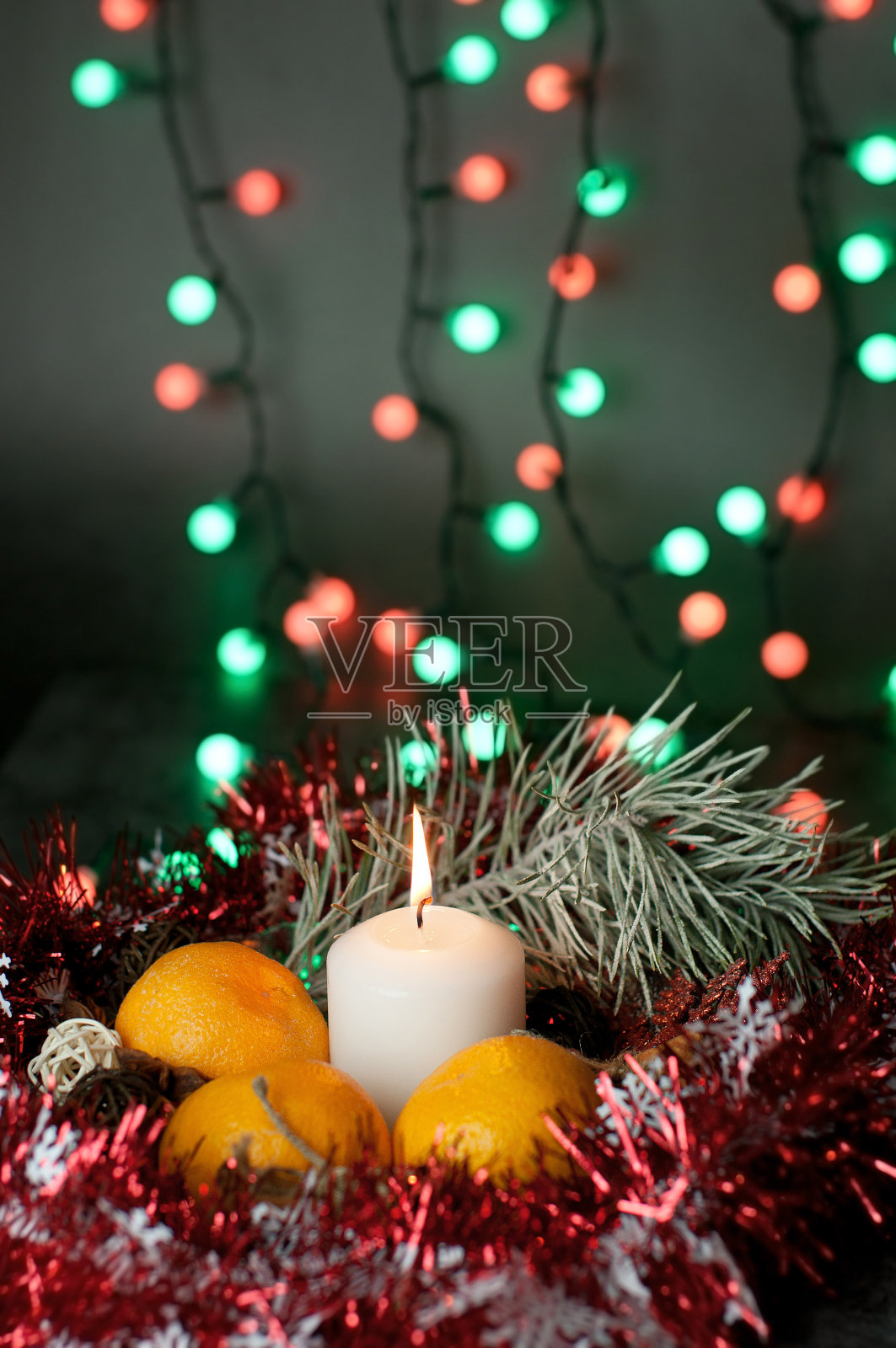 圣诞背景:桌上放着一支白色的蜡烛，上面放着牛皮纸制作的礼物、藤球、云杉花环、橘子、肉桂枝以及五彩缤纷的圣诞花环和红色雪花装饰的金属箔照片摄影图片