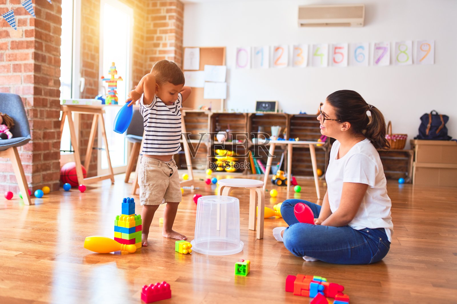 美丽的老师和蹒跚学步的男孩在幼儿园里用游戏和塑料篮子打鼓照片摄影图片