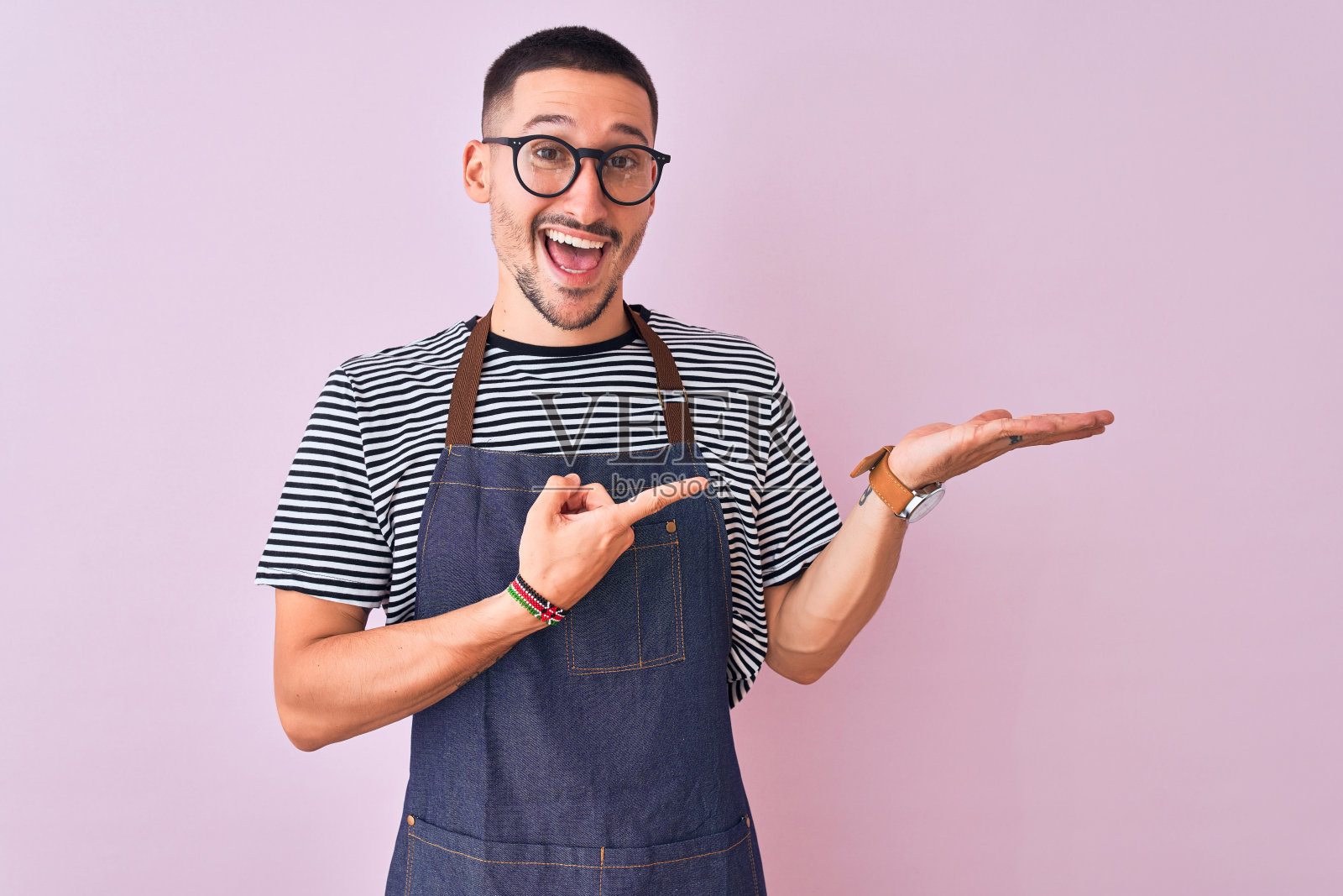 年轻英俊的男子穿着员工围裙在孤立的背景惊讶和微笑的相机，同时提出与手指。照片摄影图片