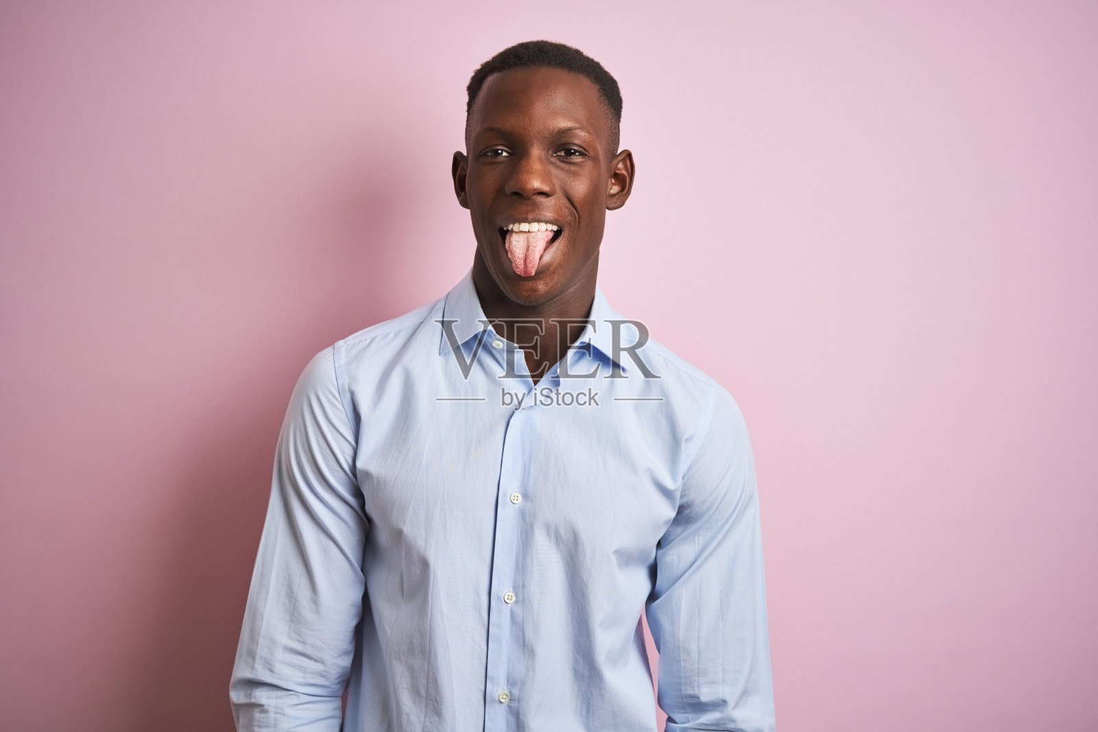 非裔美国人穿着蓝色优雅的衬衫站在孤立的粉红色背景伸出舌头快乐与有趣的表情。情感的概念。照片摄影图片