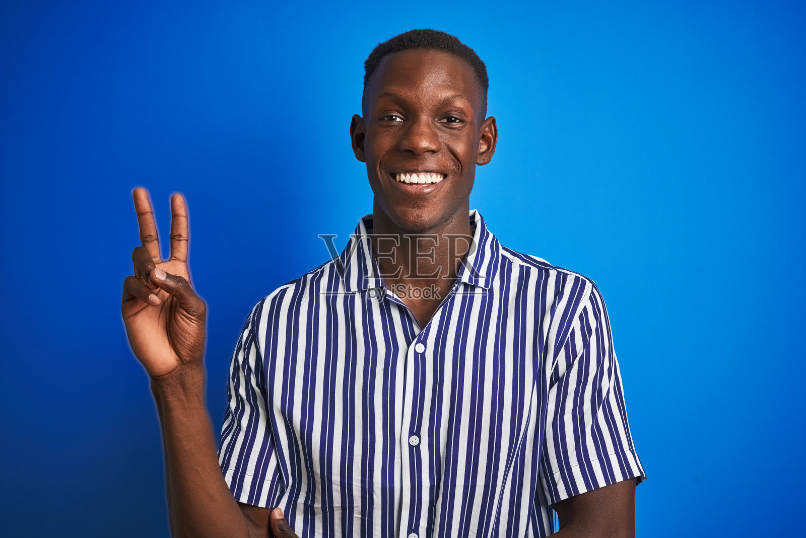 非洲裔美国人穿着条纹休闲衬衫站在孤立的蓝色背景微笑与快乐的脸在照相机眨眼做胜利标志。二号人物。照片摄影图片