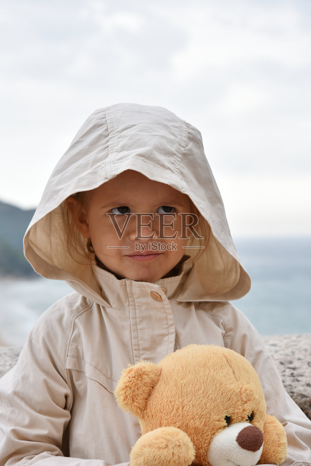 穿着带帽雨衣、抱着泰迪熊的小女孩，背景是暴风雨的天空。儿童、环境和气候概念照片摄影图片