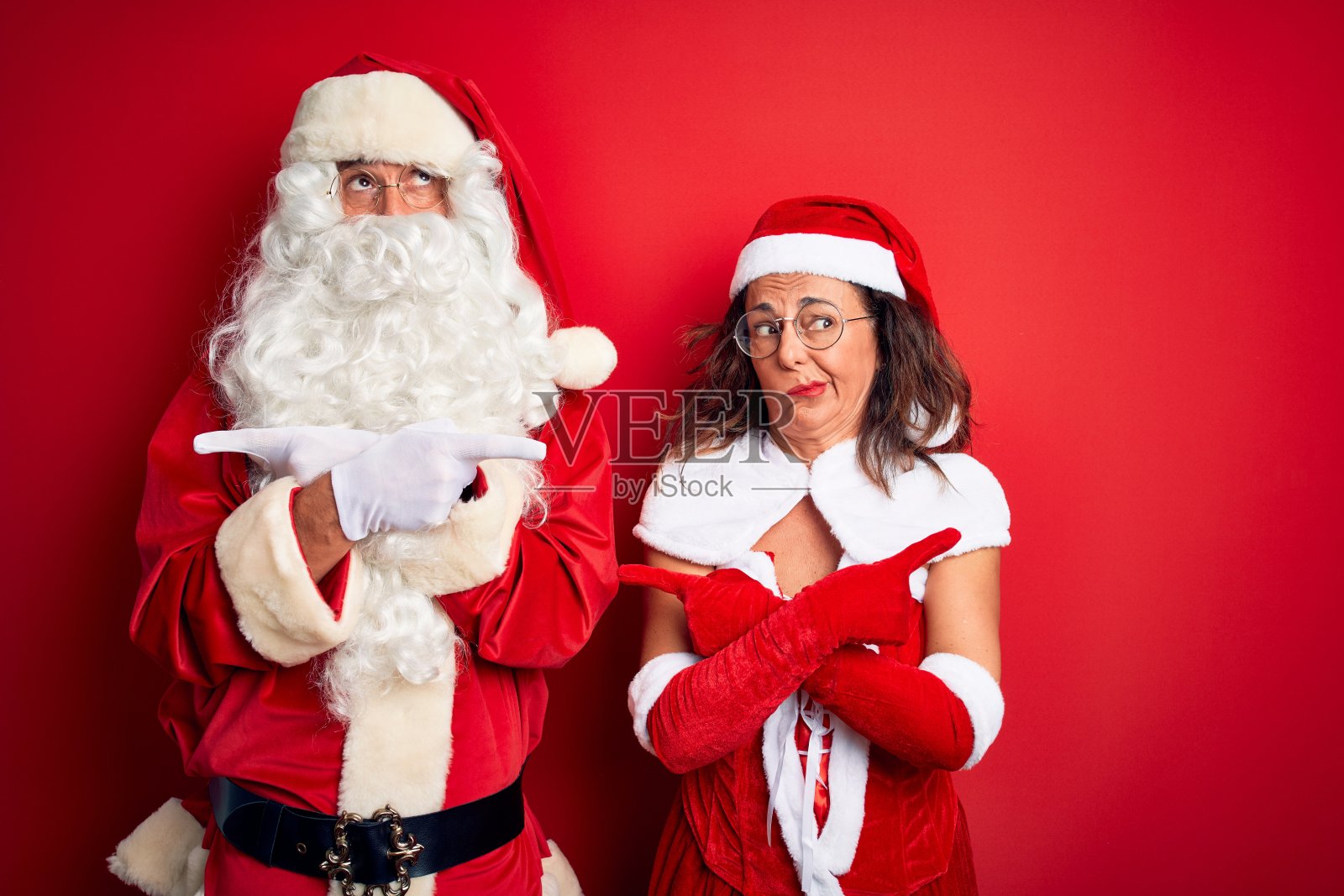 中年夫妇穿着圣诞老人服装和眼镜越过孤立的红色背景用手指指着两边，不同的方向不同意照片摄影图片