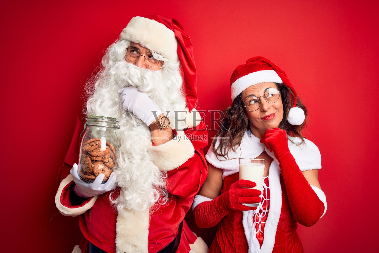 夫妇俩穿着圣诞老人服装，拿着一罐饼干和牛奶越过孤立的红色背景严肃地面对思考问题，非常困惑的想法照片摄影图片
