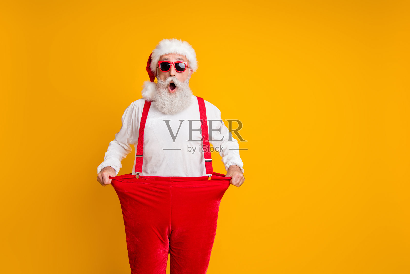 戴着圣诞老人帽的疯狂时髦的白胡子祖父拿着大尺寸的裤子，减肥，在黄色的背景下展示节食效果照片摄影图片