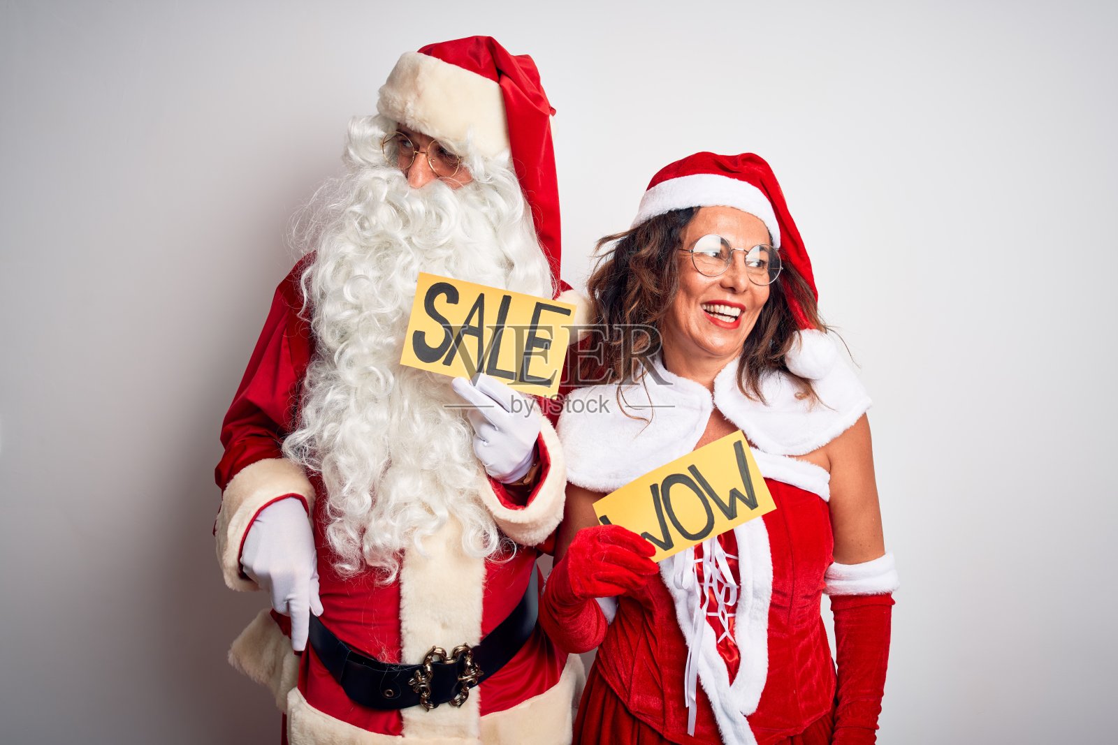 穿着圣诞老人服装的夫妇举着哇和销售旗帜在孤立的白色背景看向一边，脸上带着微笑，自然的表情。自信的笑。照片摄影图片