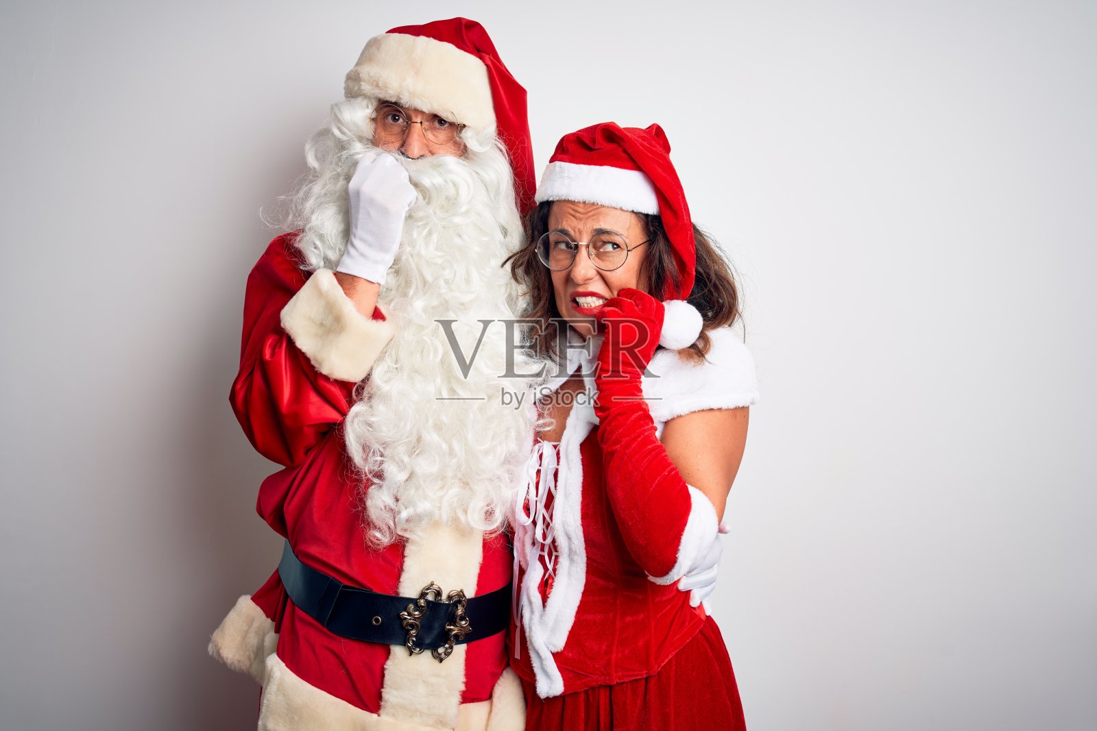 一对中年夫妇穿着圣诞老人服装，在孤立的白色背景下拥抱，看起来紧张而紧张，手放在嘴上咬指甲。焦虑的问题。照片摄影图片